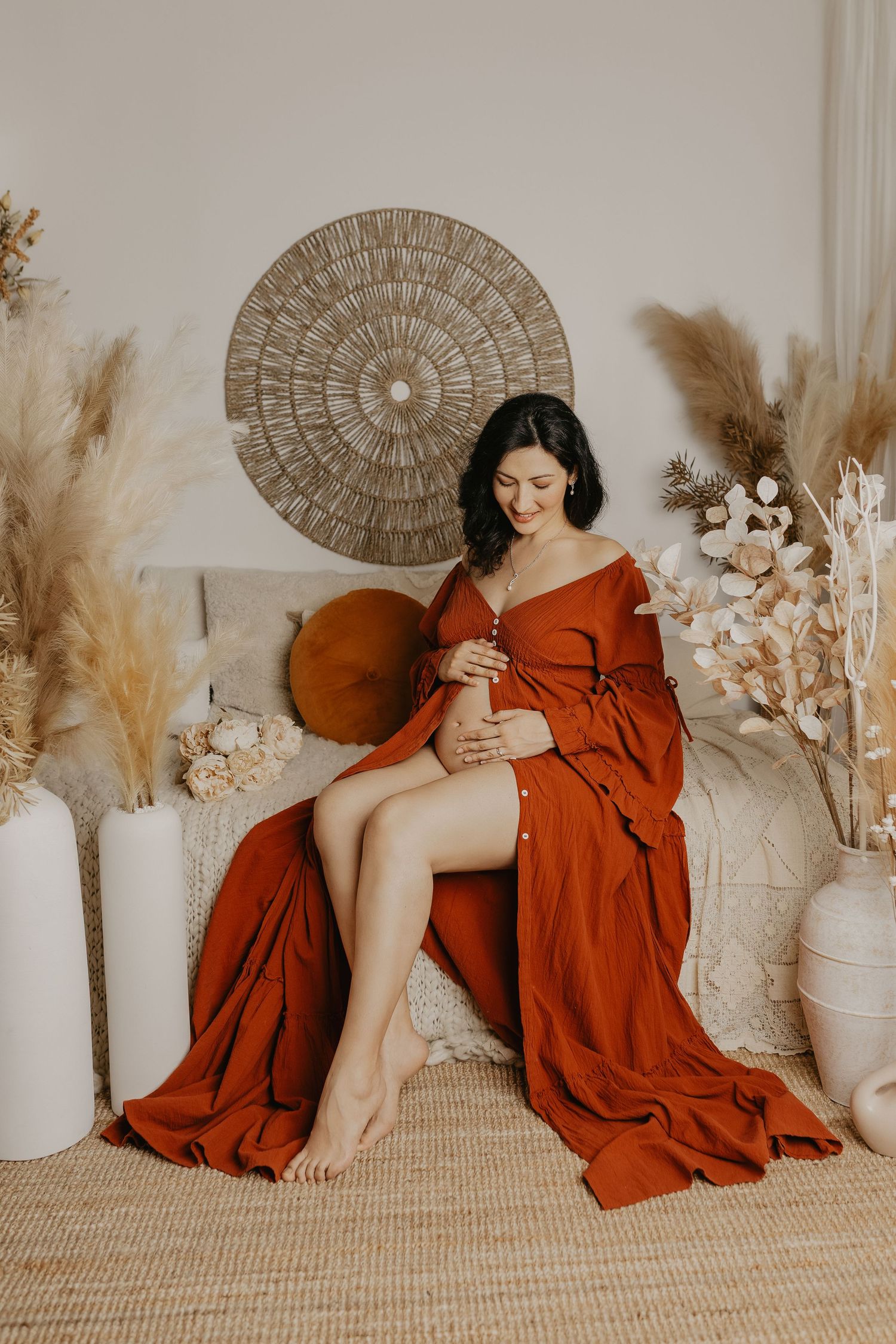 Pregnant woman in rust orange dress sitting gracefully among pampas grass decor in a boho-styled maternity photo shoot.