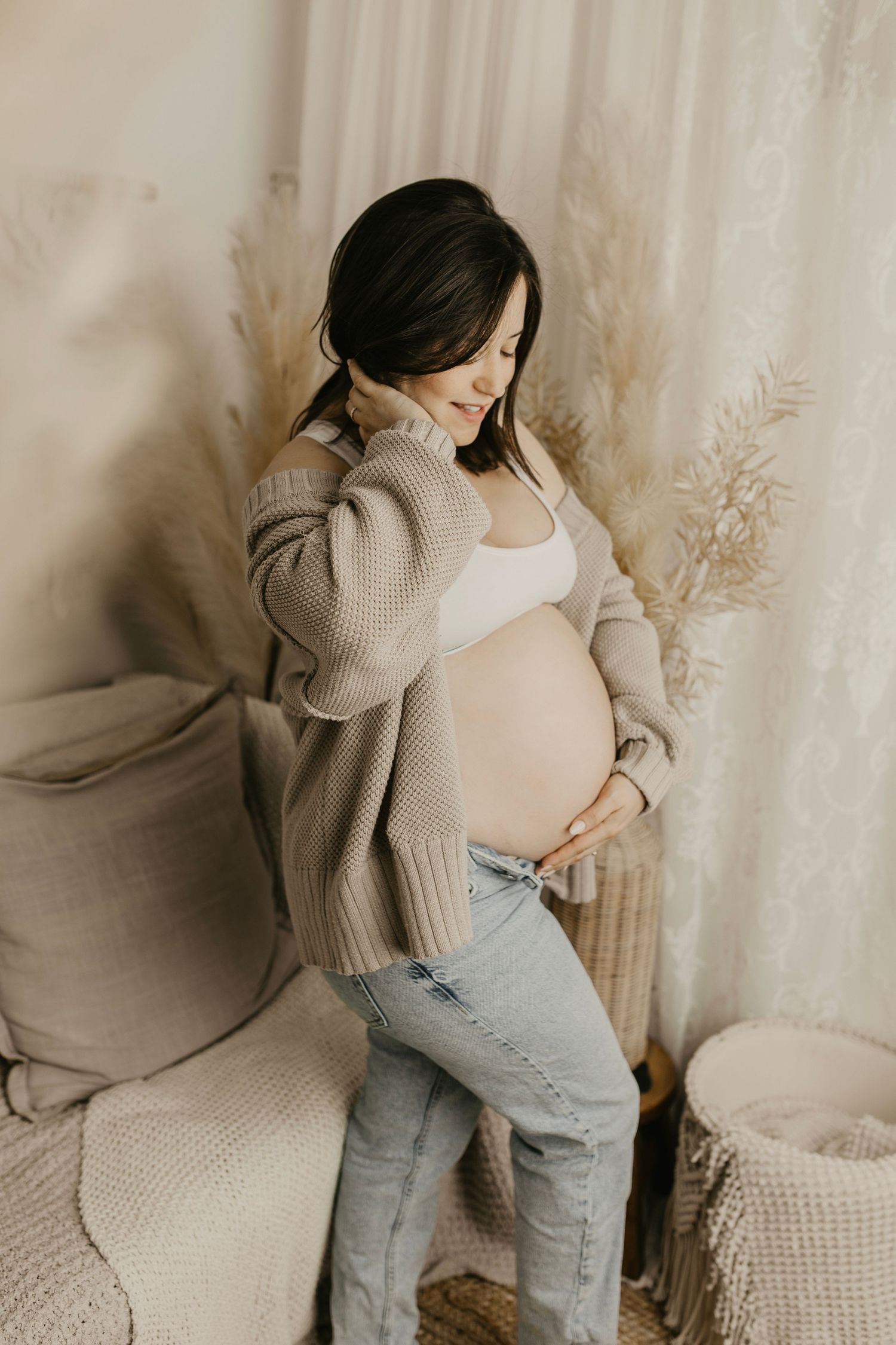 Expectant mother in beige sweater and jeans poses against neutral backdrop.