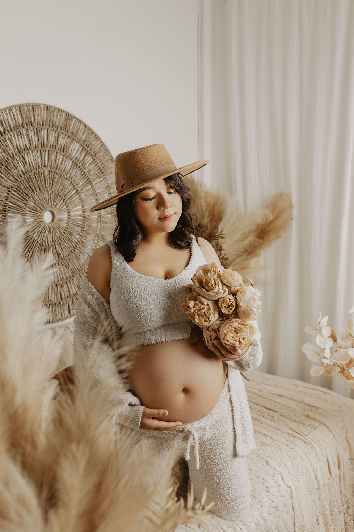 Pregnant woman in boho style holding dried flowers, wearing tan hat and white crop top, surrounded by pampas grass decor.