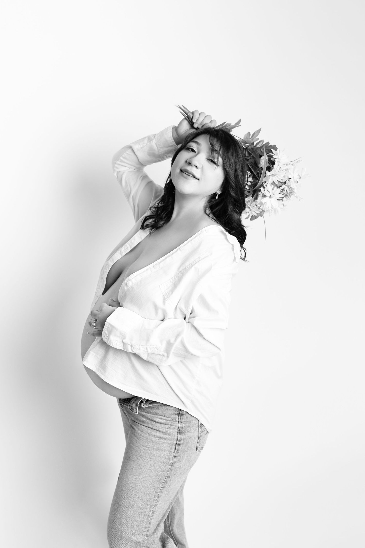 Black and white maternity photo of a woman in jeans and white top wearing a floral crown, posing gracefully.
