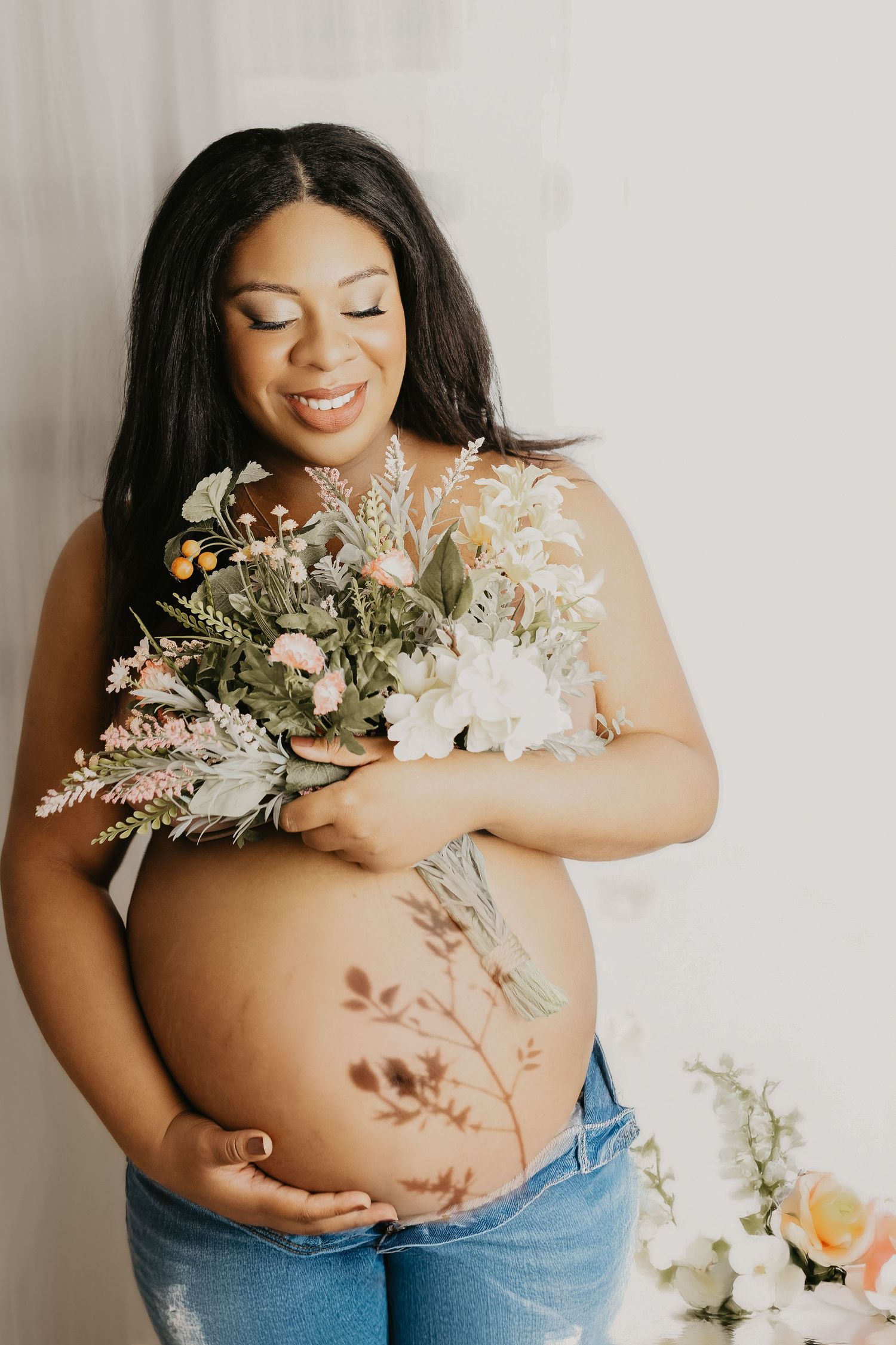 Artistic maternity photoshoot featuring floral arrangements and a casual denim style against a light background.
