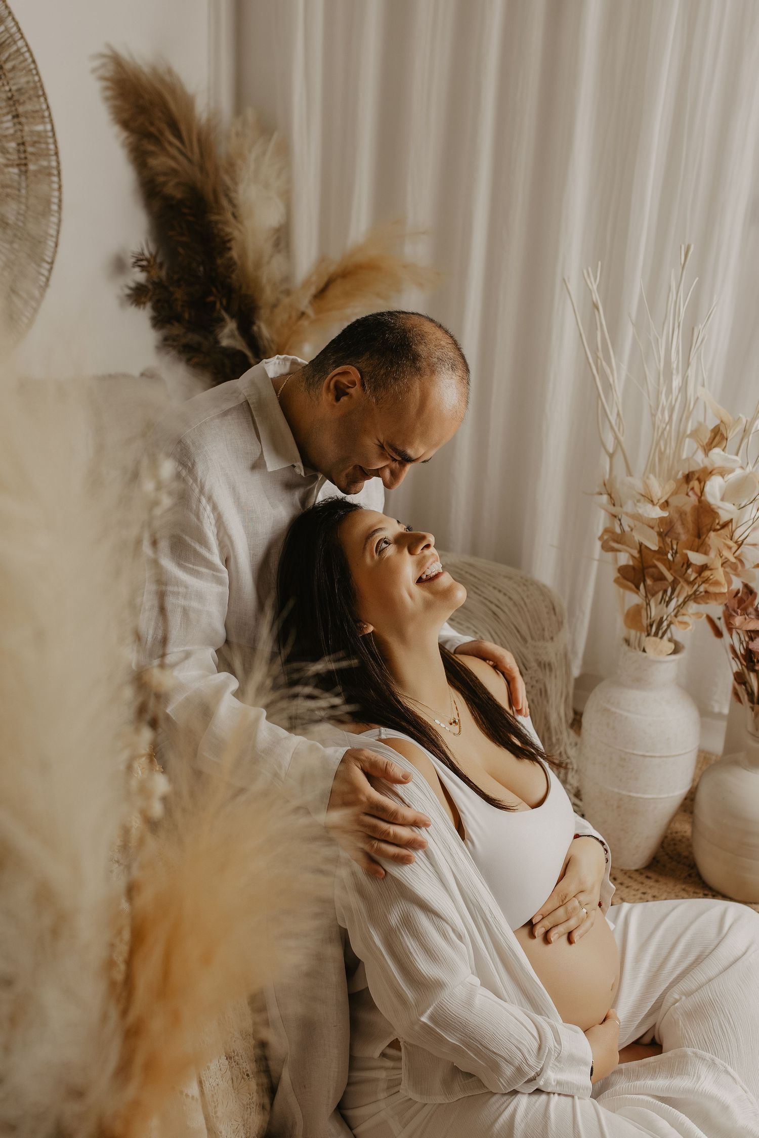 A couple shares a tender moment, with a man embracing a pregnant woman surrounded by boho dried floral decor.