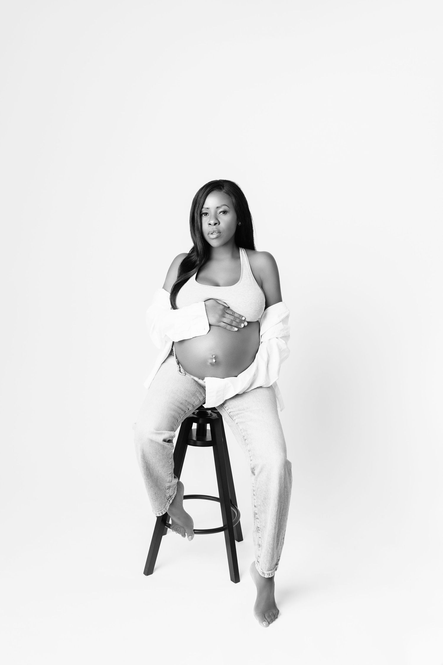 Black and white maternity photo of a pregnant woman sitting on a stool, wearing ripped jeans and a crop top.