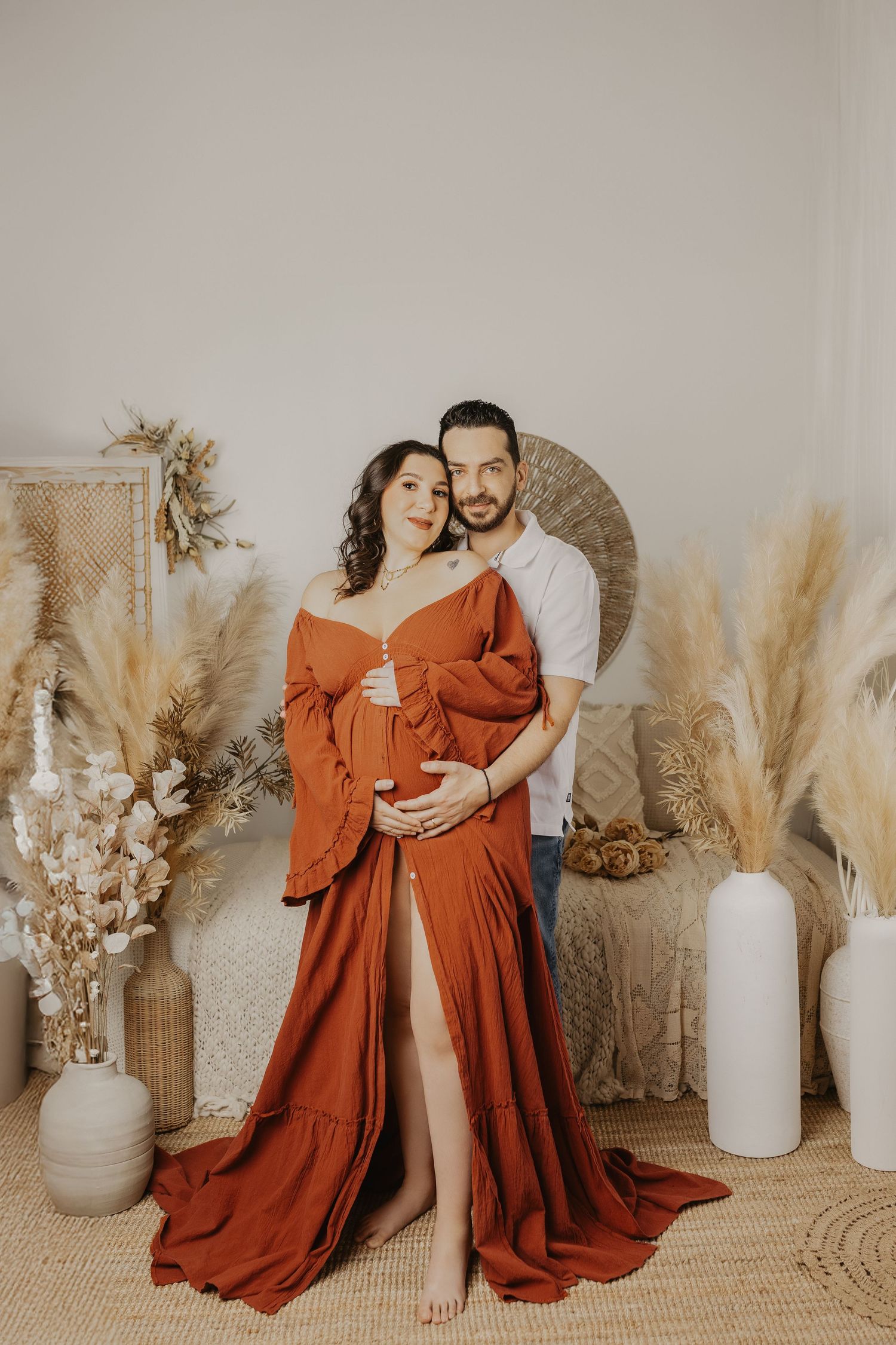 A romantic maternity photoshoot with a flowing rust-colored dress against a neutral backdrop with dried pampas grass decor.