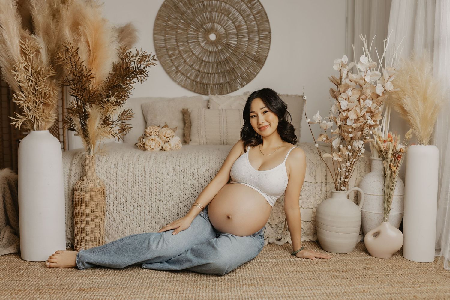 A maternity photo featuring a boho-style setup with neutral decor, dried pampas grass, and ceramic vases on a beige carpet.