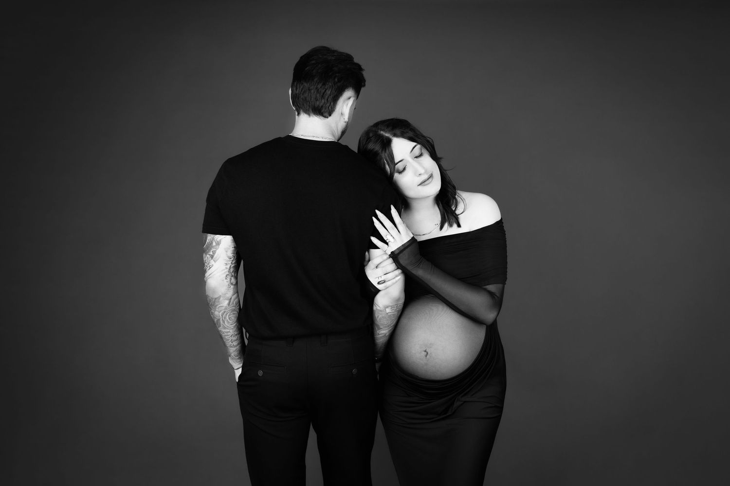 Black and white maternity photo of pregnant woman leaning on partner's shoulder in studio setting.