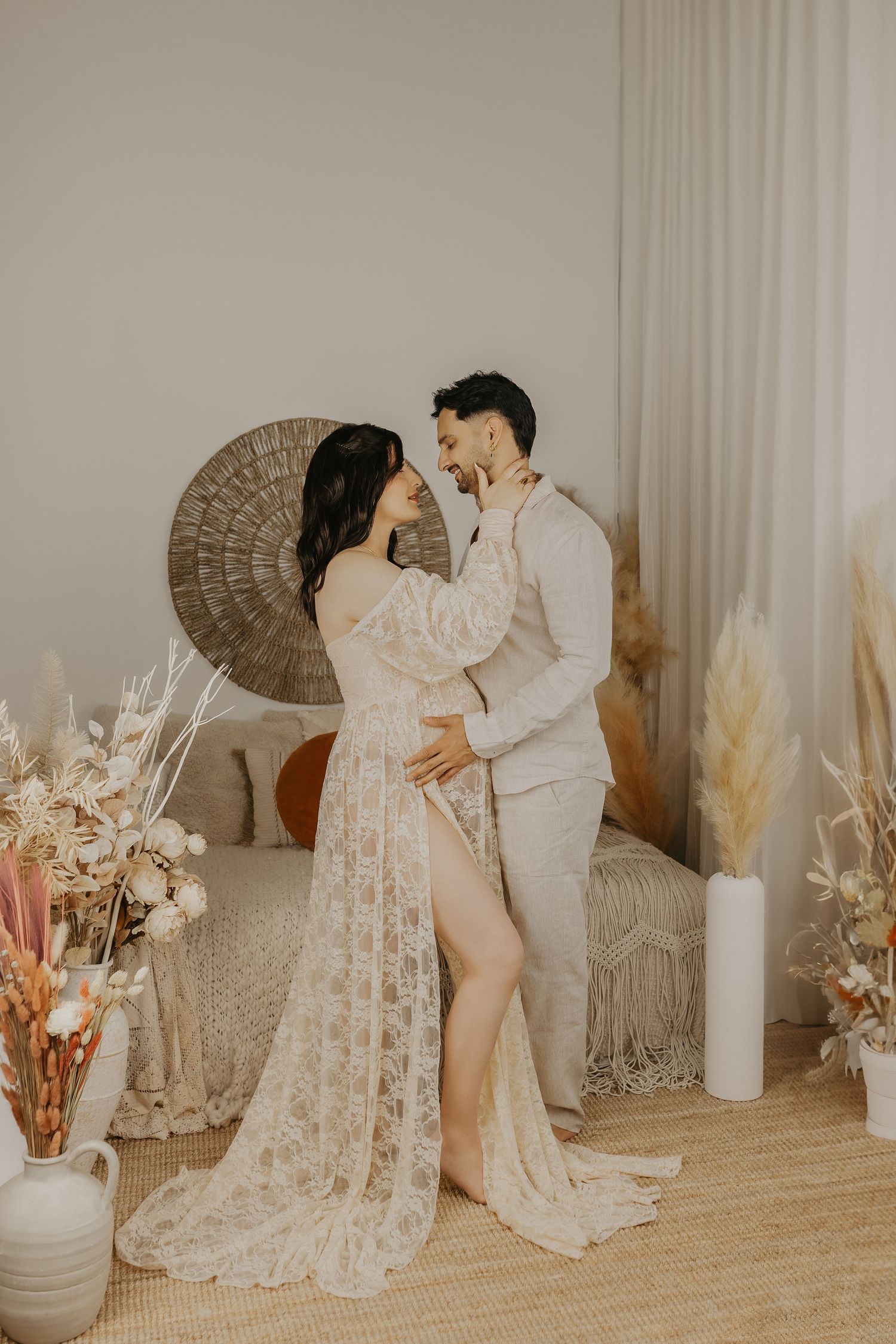 Couple in boho wedding attire embracing in a neutral-toned room with pampas grass and dried floral arrangements.