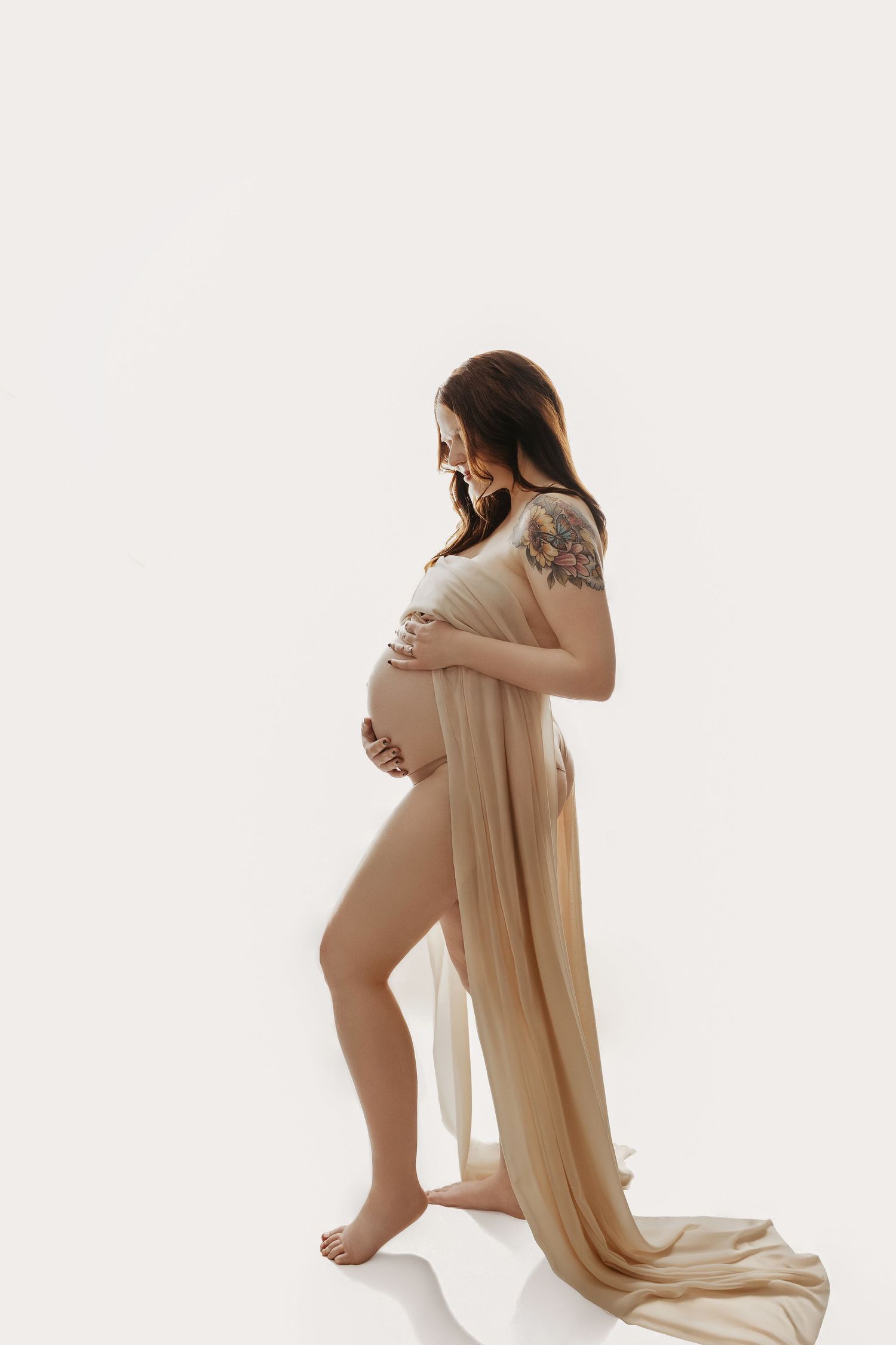Artistic nude maternity silhouettes draped in flowing beige fabric against white background.