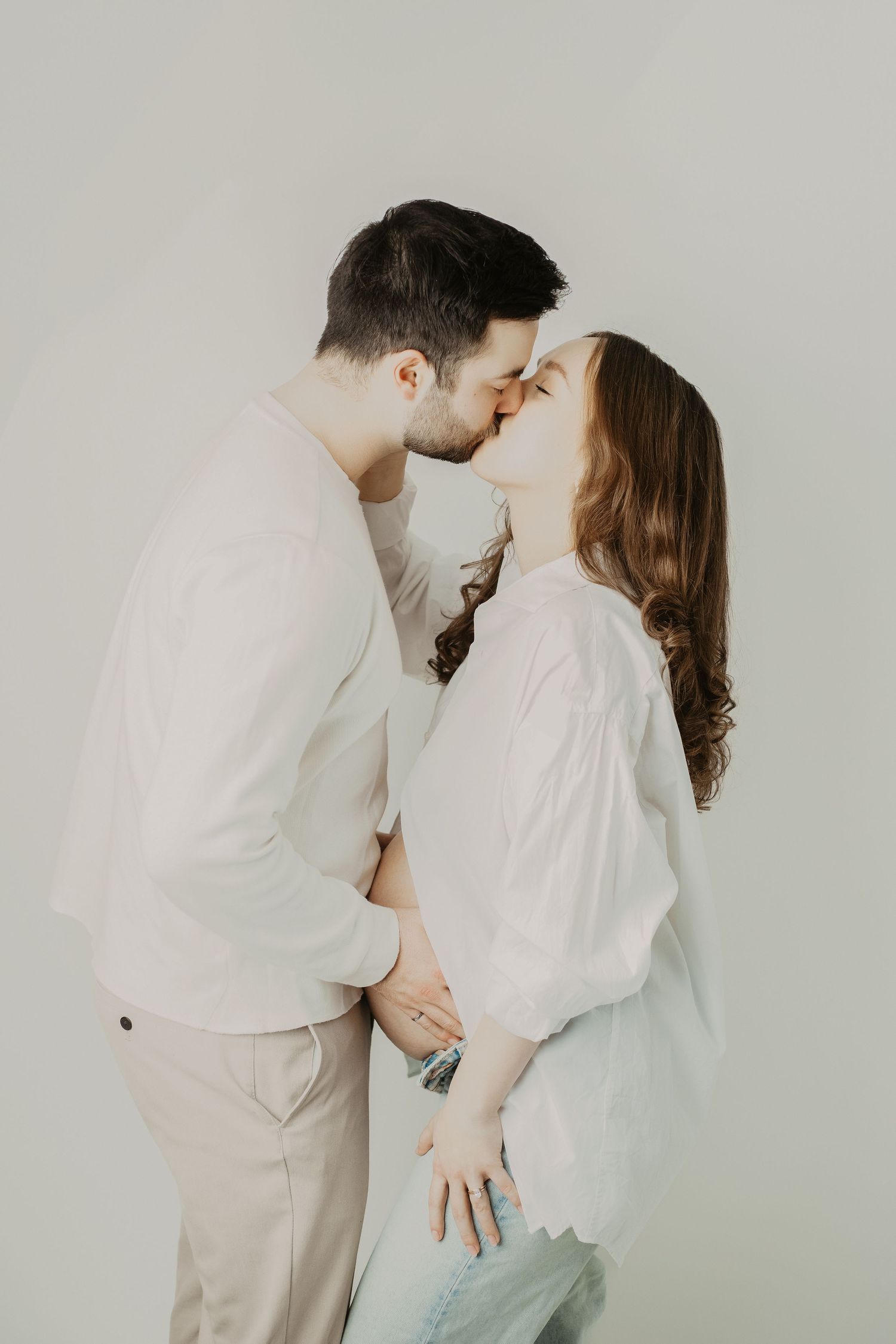 Intimate maternity portrait of couple in white sharing a tender moment against neutral background.
