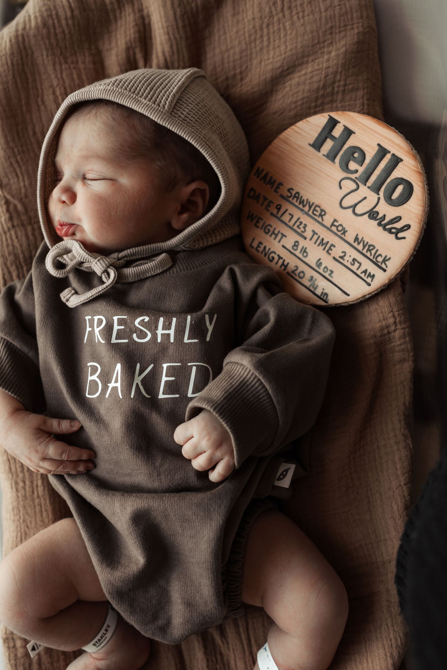 Newborn baby wearing brown hooded 'Freshly Baked' onesie next to birth announcement sign.