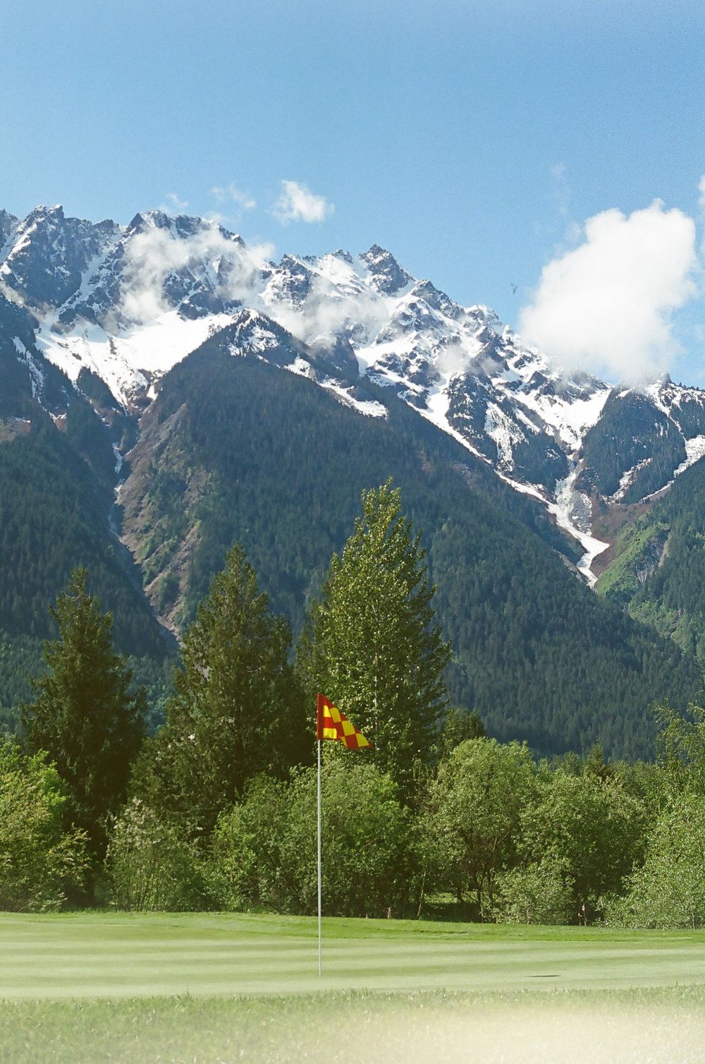 Golf course with yellow flag against dramatic snow-capped mountain landscape.