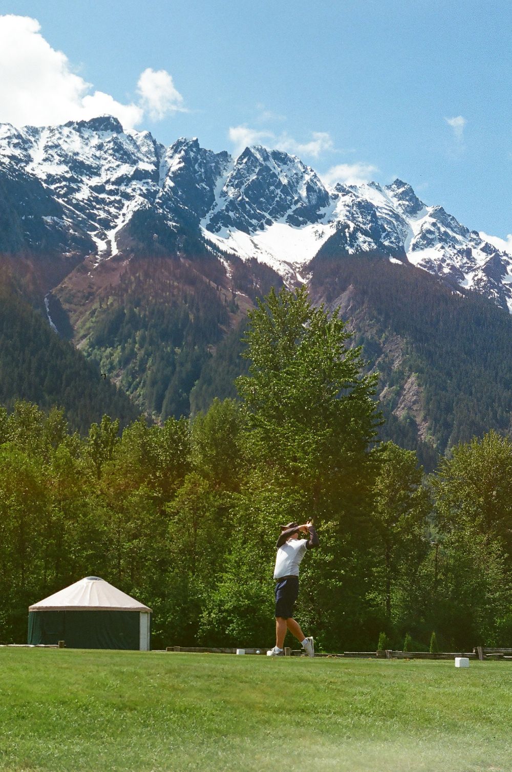 Golf course with white tent and players set against towering snow-covered mountain peaks.