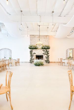 Modern white wedding venue with wooden chairs and a decorated brick fireplace focal point.