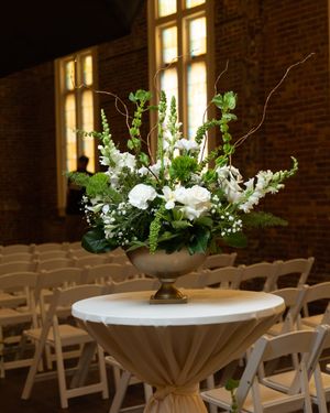 Elegant white and green floral arrangement in a gold pedestal vase on a cocktail table with folding chairs in the background.