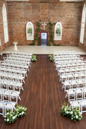 Church ceremony setup with white chairs, floral arrangements, and a wooden cross at the altar.