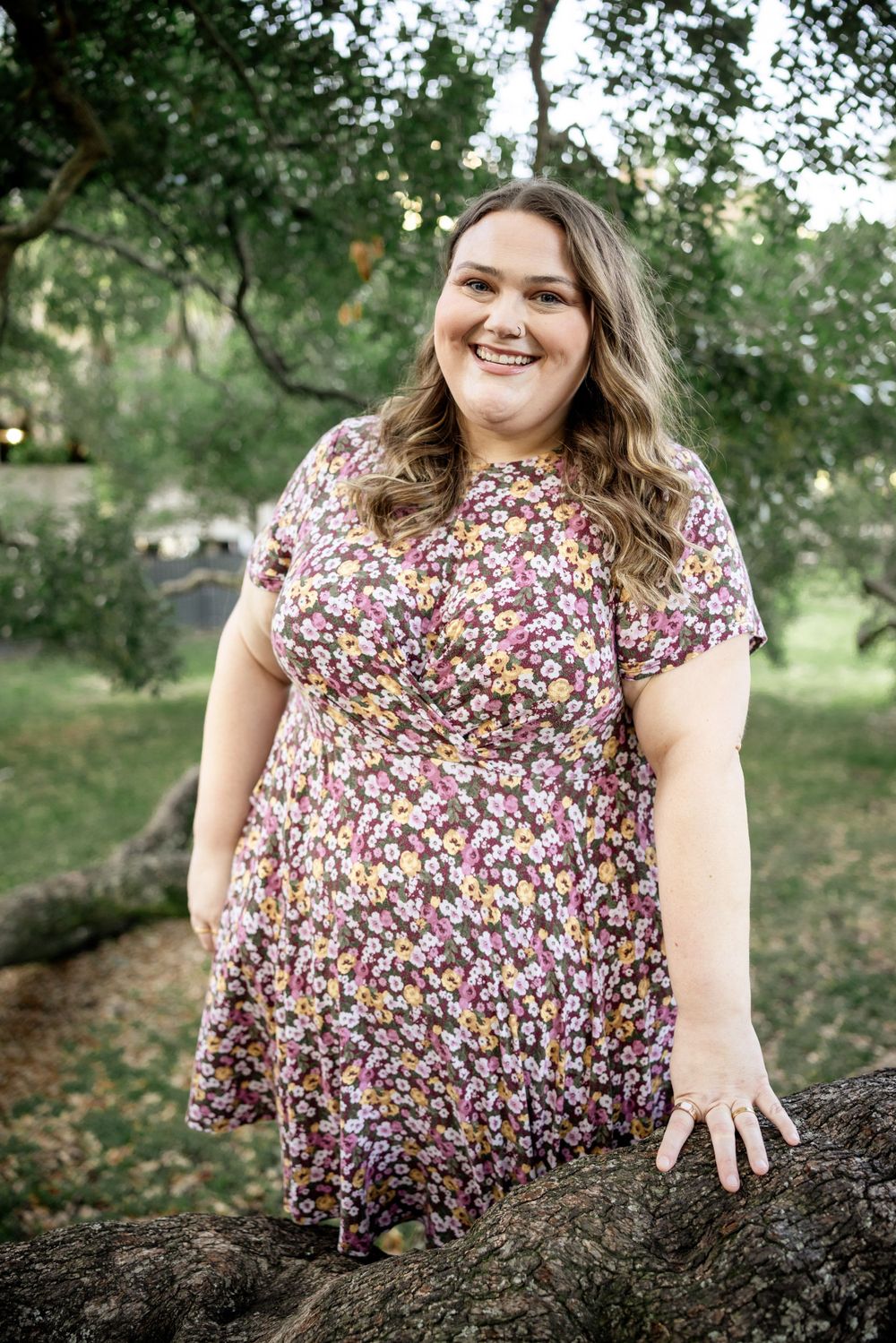 Woman in floral dress sits on tree branch outdoors with genuine smile.