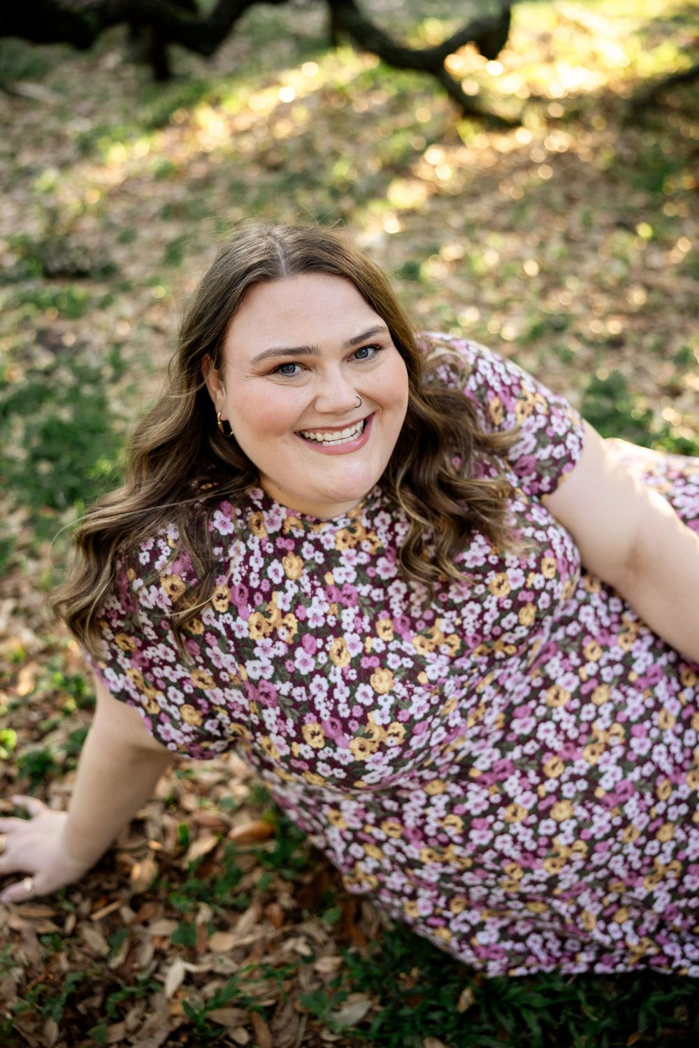 Woman in floral dress smiling while sitting on grass in outdoor garden setting.