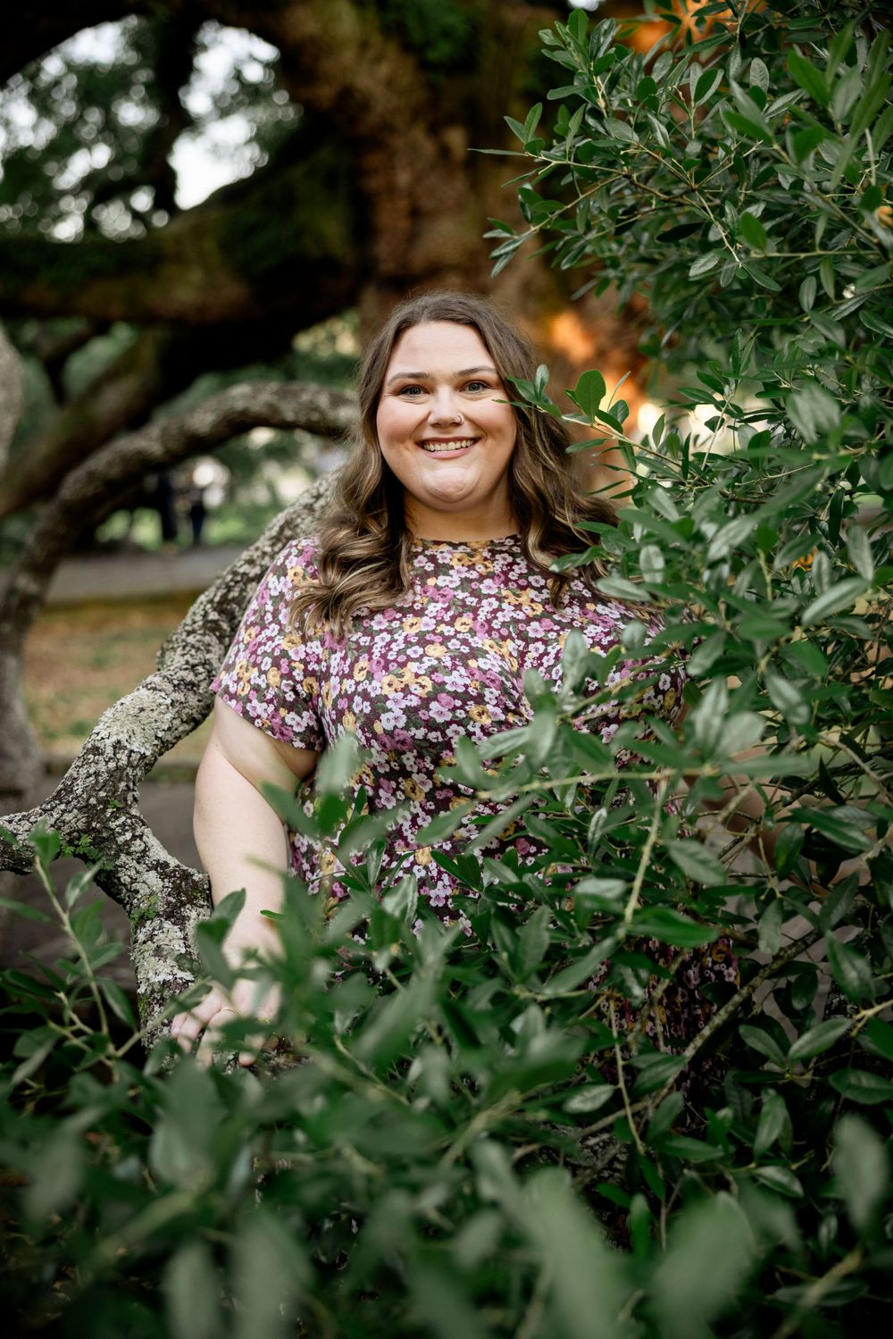 Woman wearing floral dress poses among green garden foliage with natural lighting.