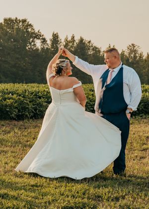 Groom with one hand in his pocket twirls his elegantly dressed bride in a field at sunset during golden hour.