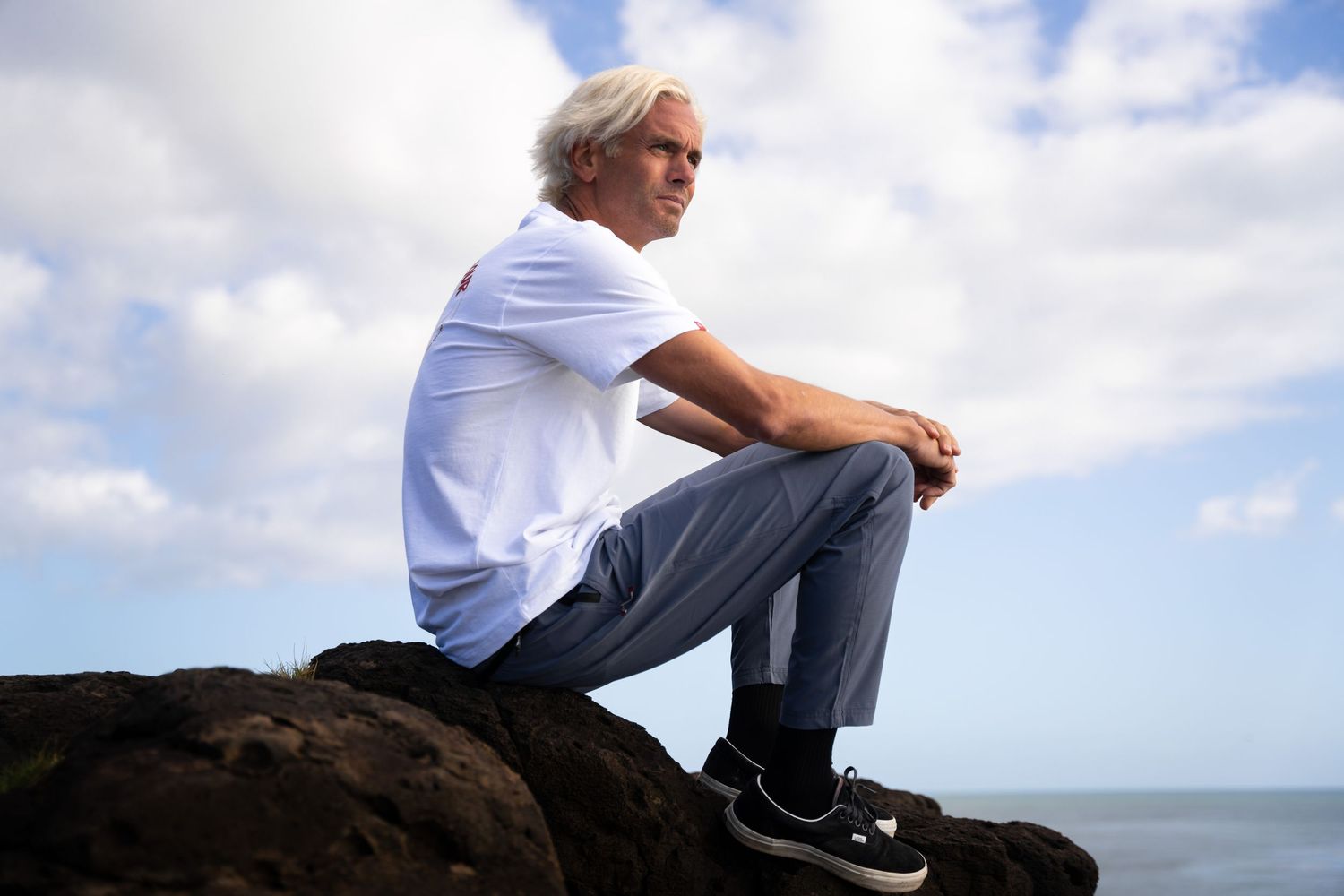 Person in white shirt and gray pants sits contemplatively on rocky coastal outcrop with ocean and cloudy sky in background.
