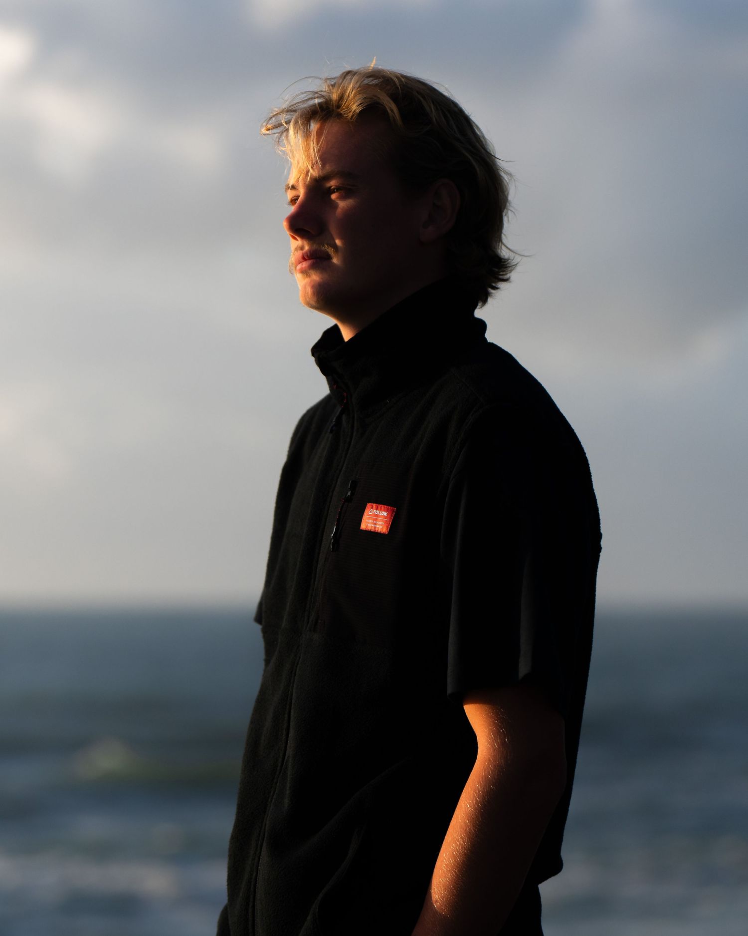 Silhouetted figure stands against dramatic cloudy sky and ocean horizon at sunset.