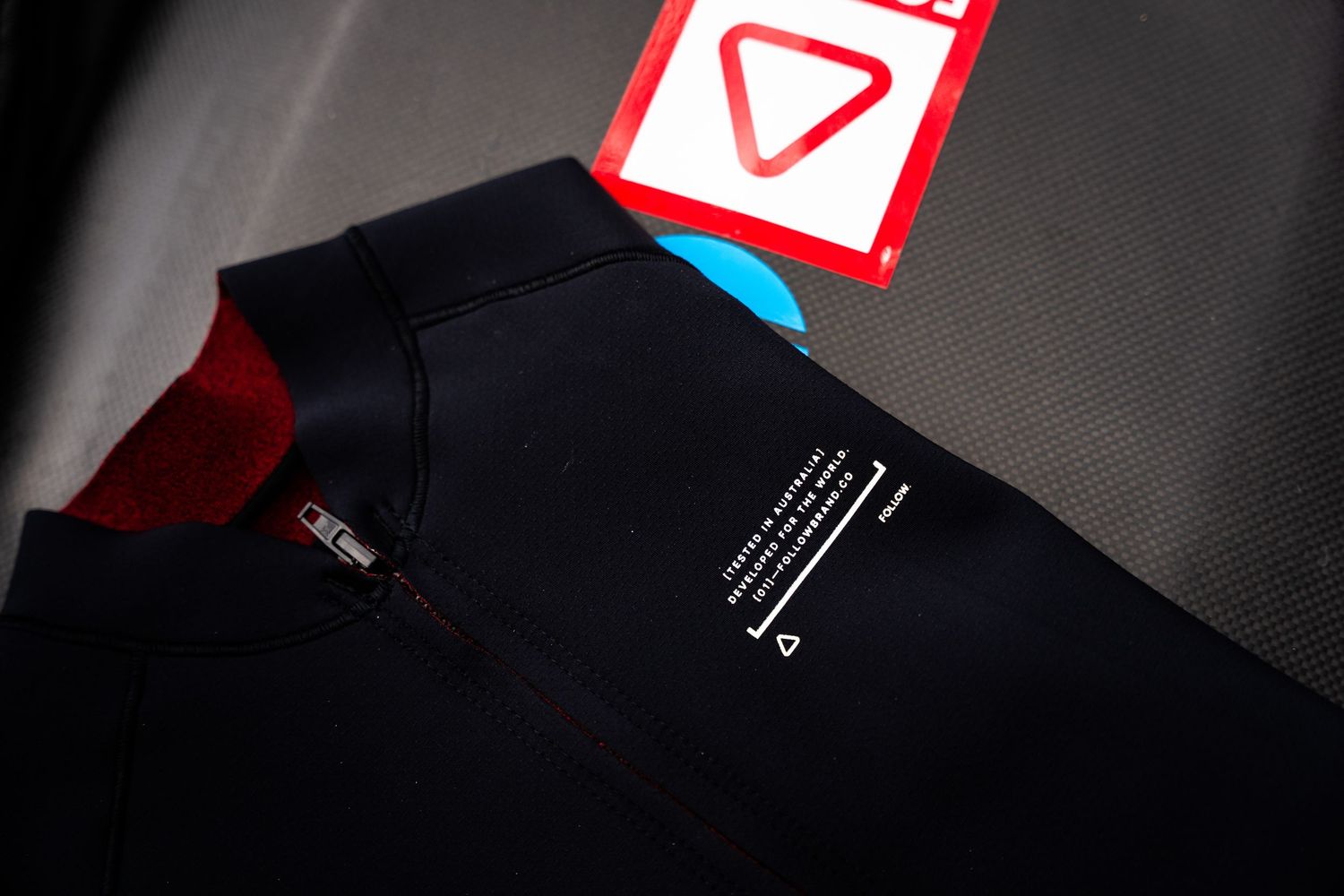 Close-up view of a black technical garment with a red tag and printed text details on dark background.