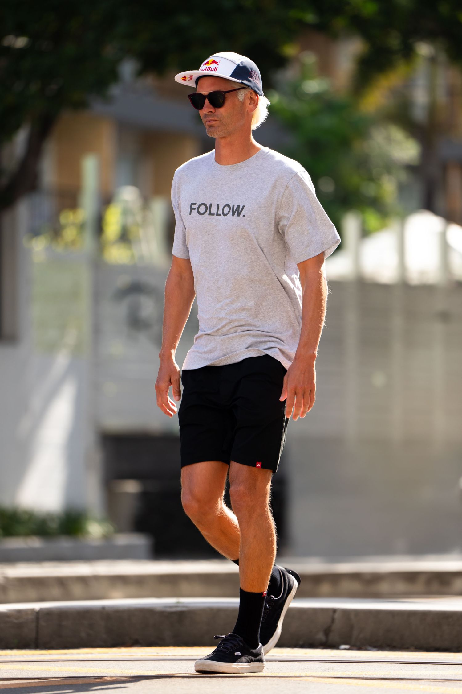 A person wearing sunglasses, a baseball cap, white t-shirt with 'FOLLOW' text, and black shorts walks outdoors on a sunny day.