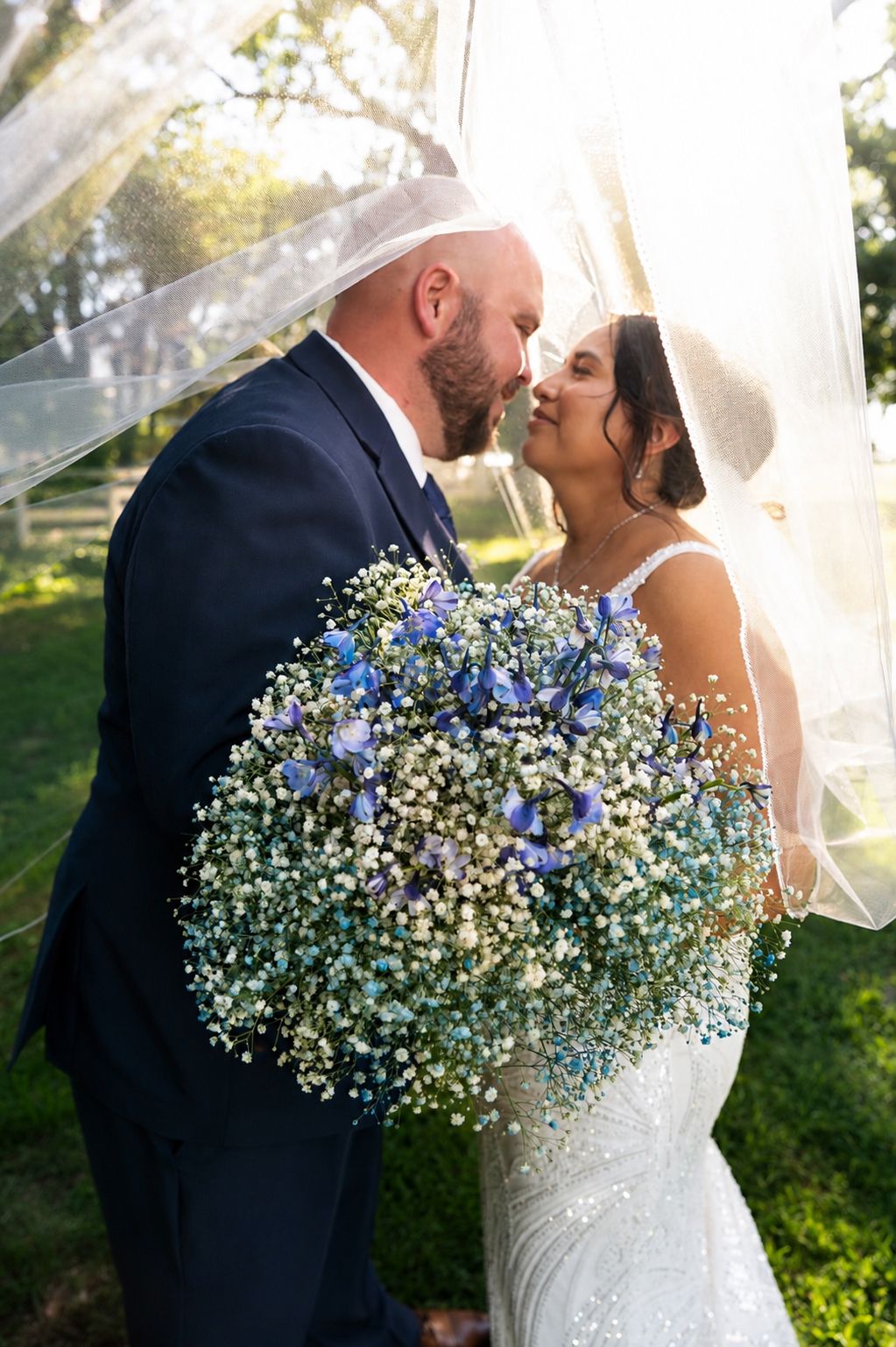 Bride and groom share a romantic kiss under a flowing veil, holding a blue and white baby's breath bouquet outdoors.