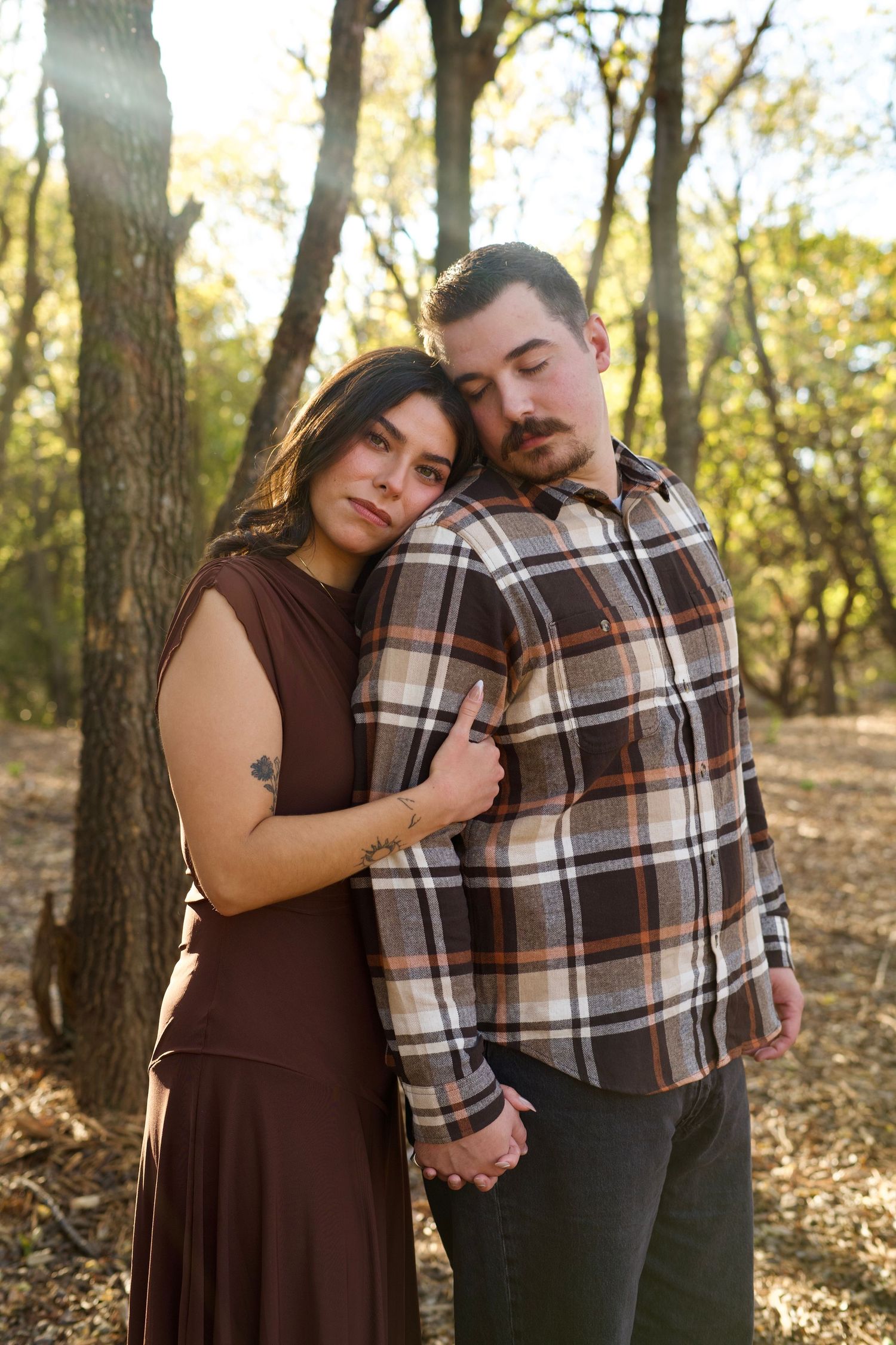 Couple in autumn forest embracing, wearing brown dress and plaid shirt in warm sunlit woodland setting.