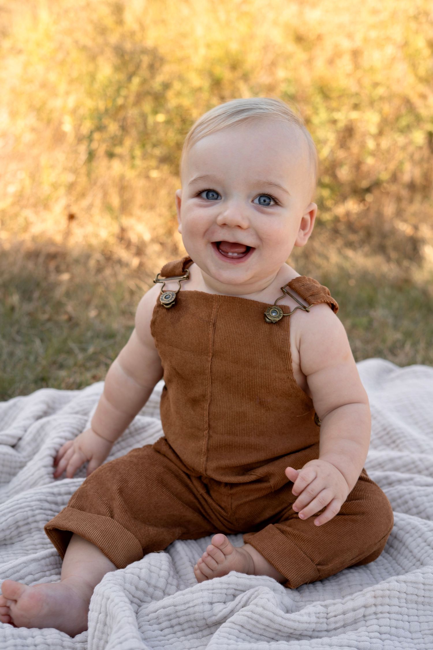 A smiling baby in brown corduroy overalls sits on a textured gray blanket in a sunny outdoor setting.