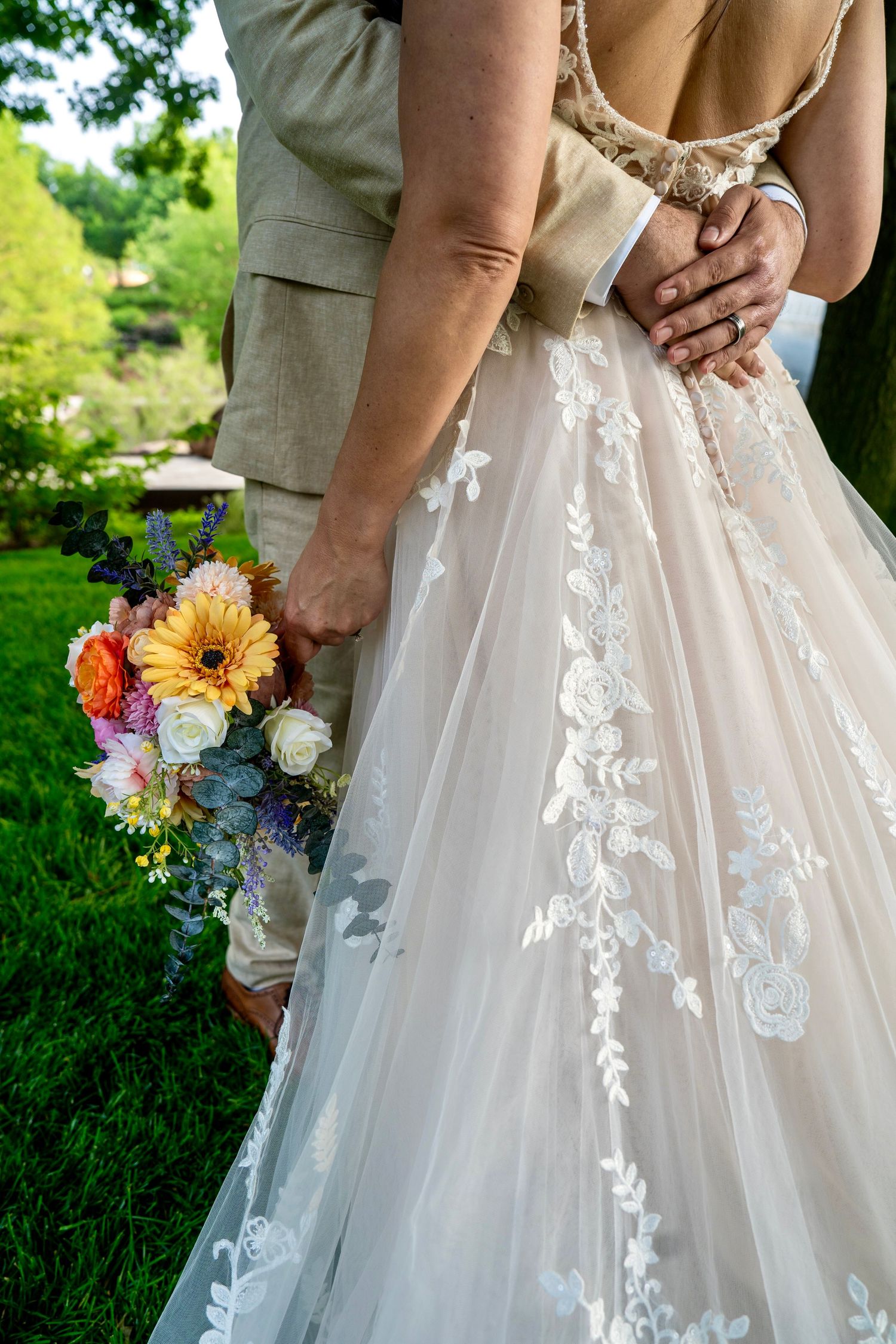 Bride in lace wedding gown holding colorful bouquet with groom in tan suit, showing wedding rings outdoors.