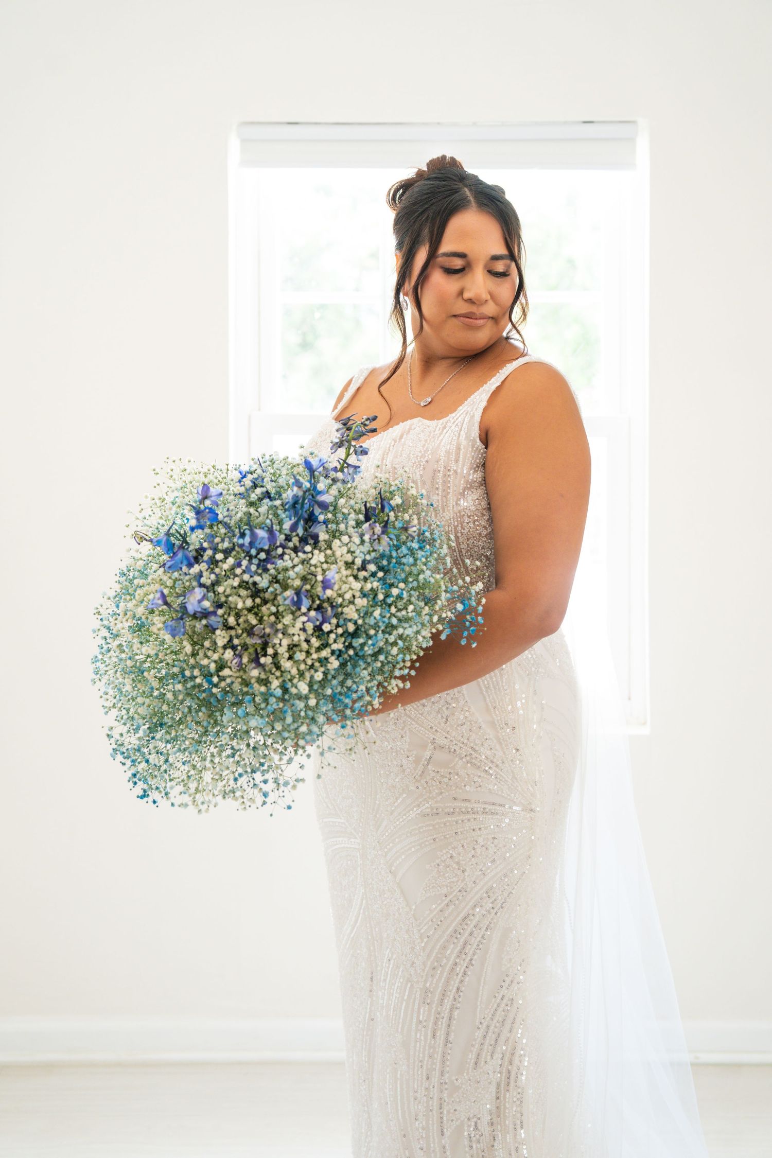 Bride in beaded white gown holding large blue and white baby's breath bouquet, standing before bright window.