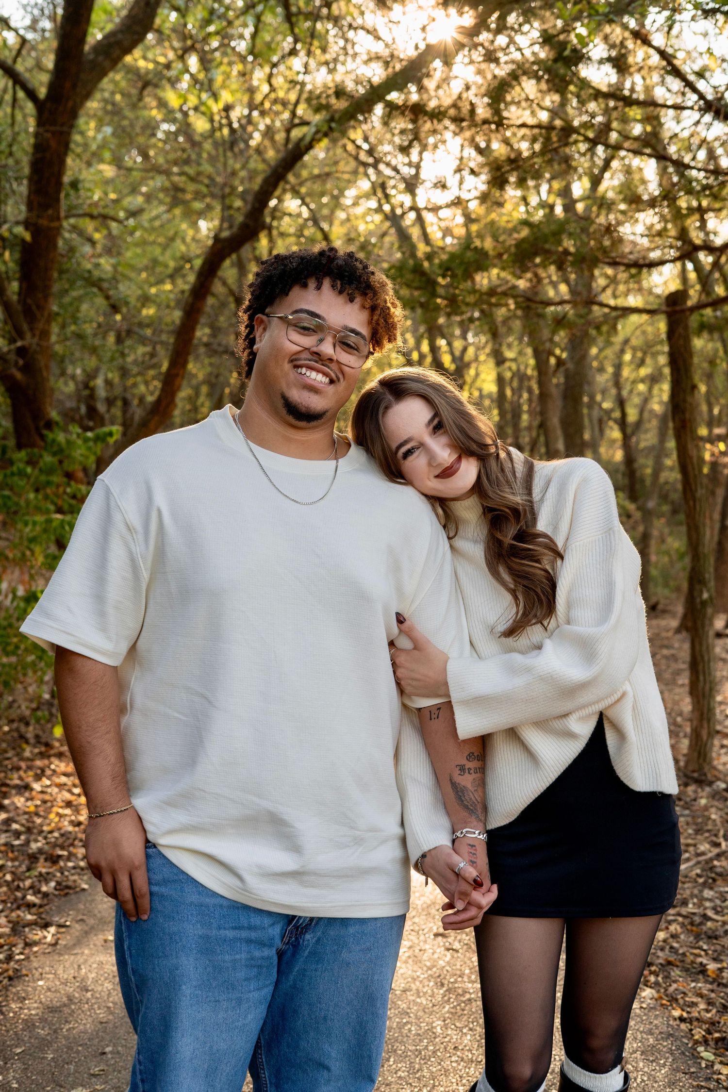 Couple in white tops and denim embrace while standing in a sunlit wooded area during golden hour.