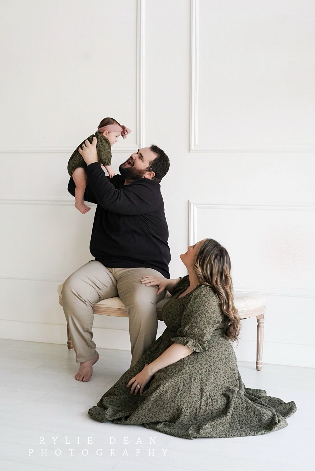 Indoor family portrait with parent lifting baby while seated on bench as another figure in green dress sits nearby.