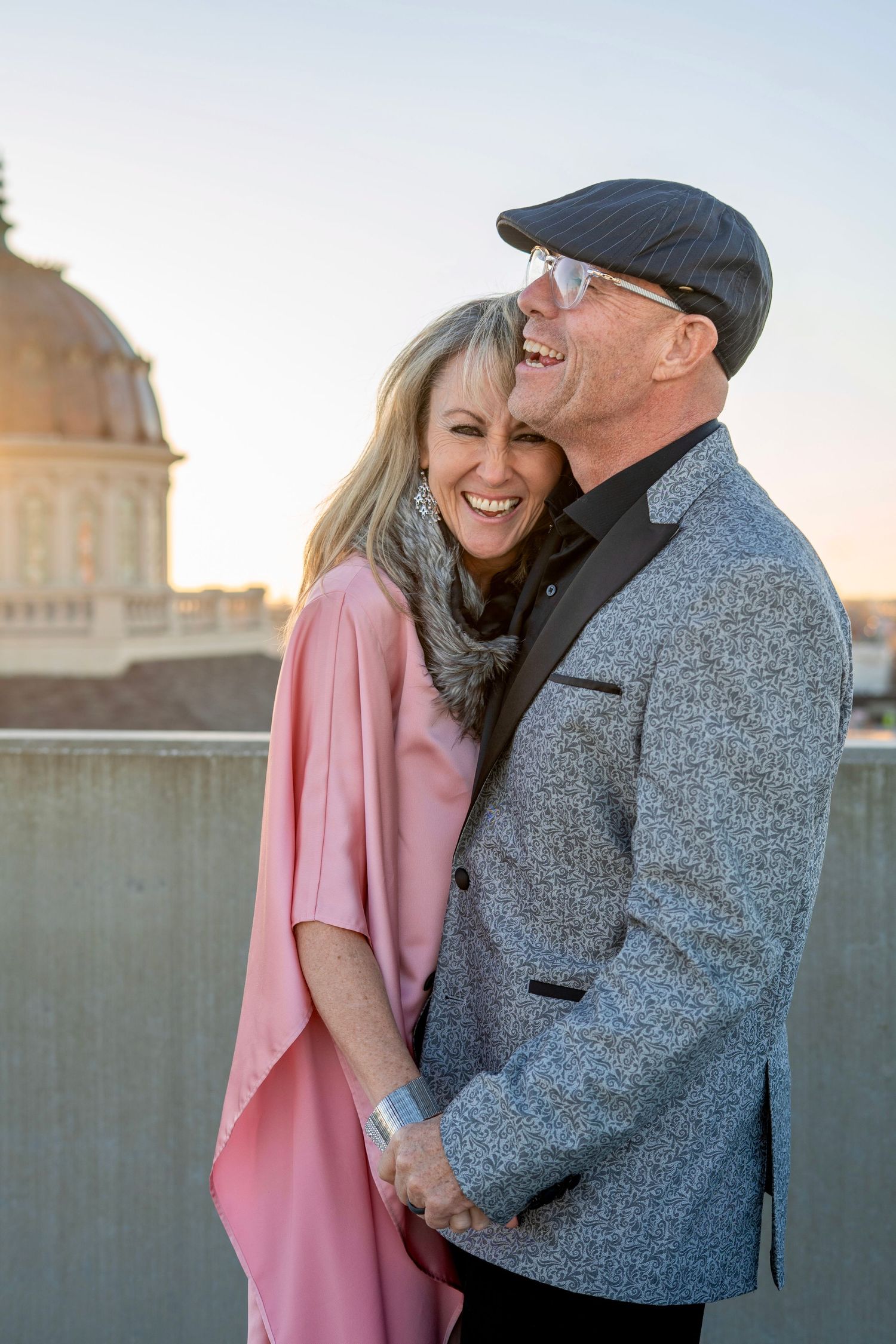 Happy couple laughing together outdoors at sunset, wearing pink dress and grey blazer with flat cap.
