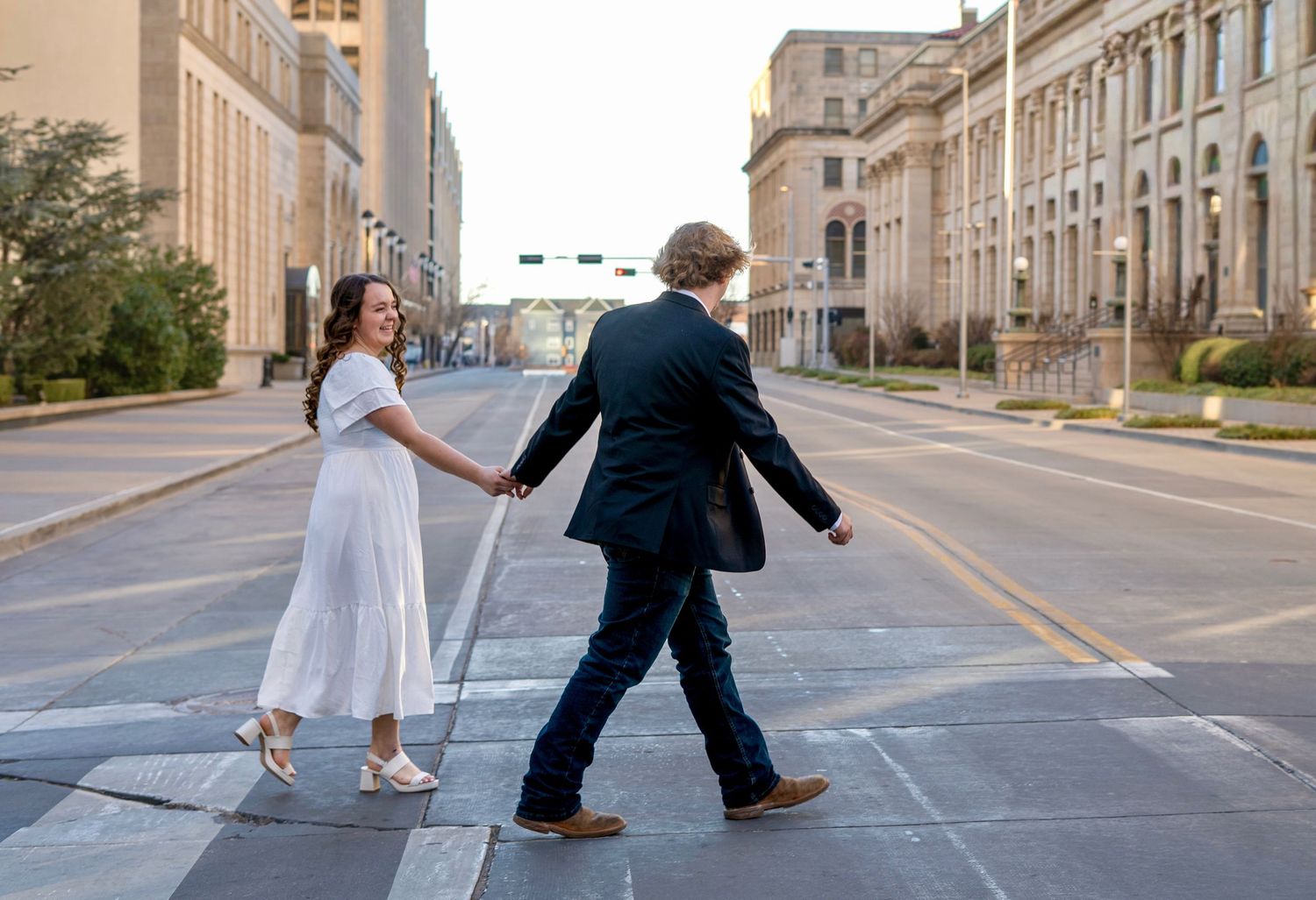 A couple in formal attire walks down the middle of an empty city street with historic buildings on both sides.