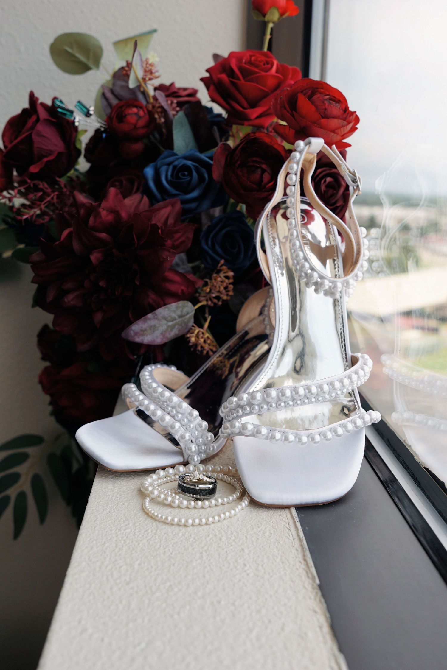 Elegant white bridal shoes displayed beside deep red roses and a pearl necklace on a windowsill.
