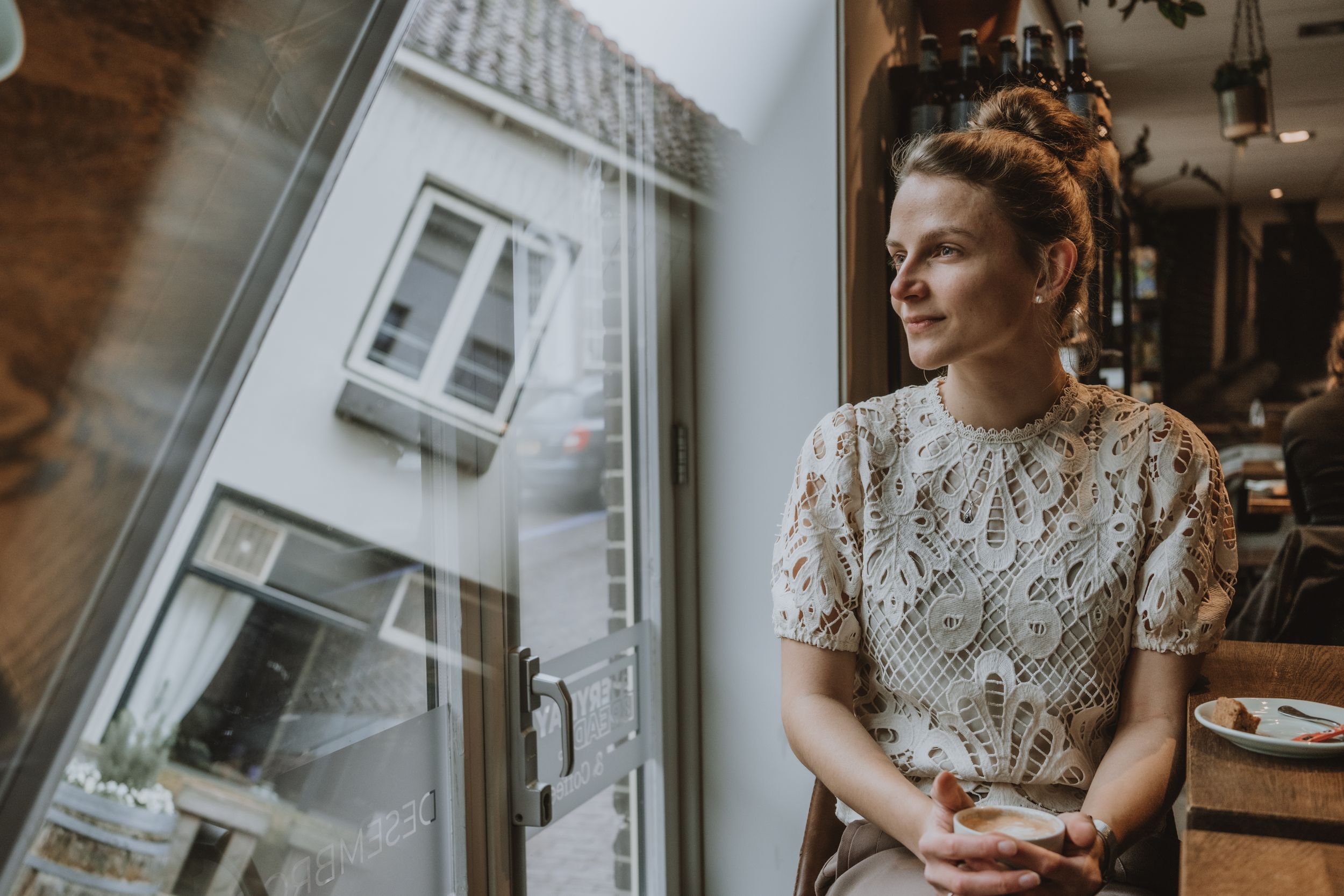 Young woman in lace dress gazing thoughtfully out a window in a cozy cafe setting.