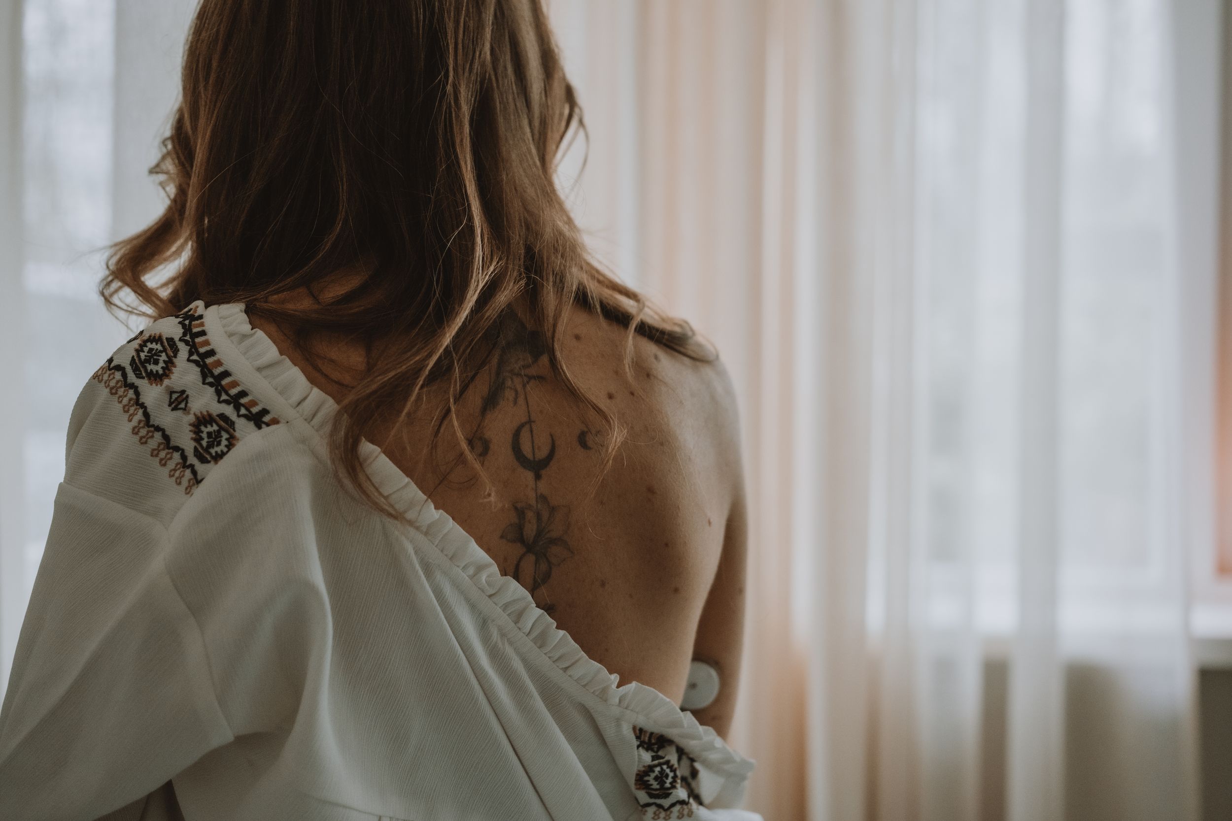 Close-up of a woman showing celestial tattoos on her upper back near a bright window.