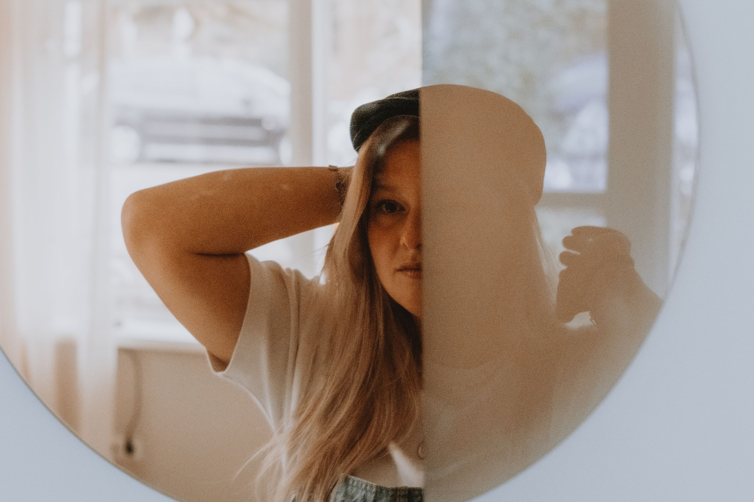 Blonde woman with hands in hair reflected in a round mirror near a bright window.