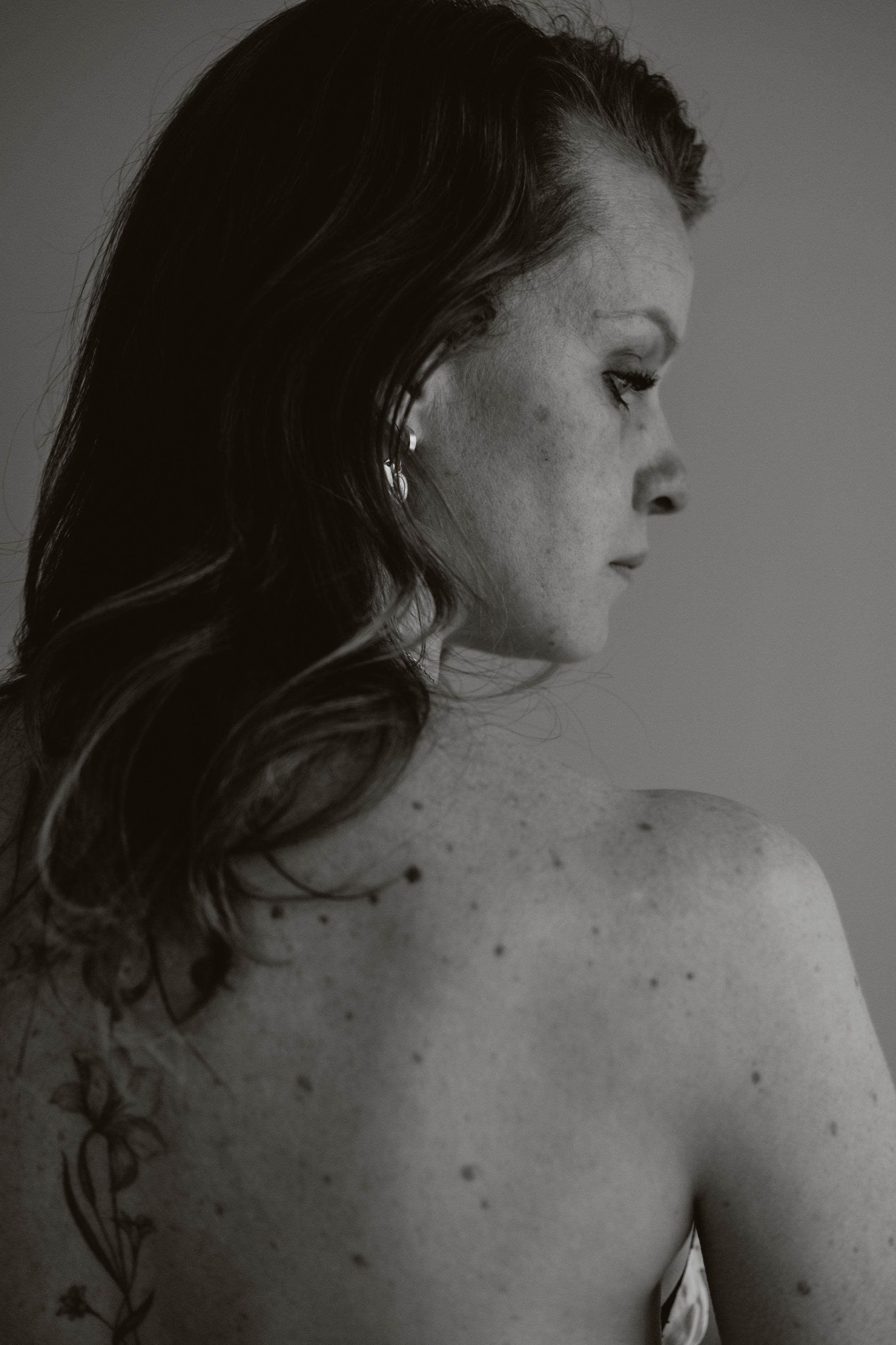 Black and white portrait of a freckled woman with dark hair showing her bare back with a tattoo.