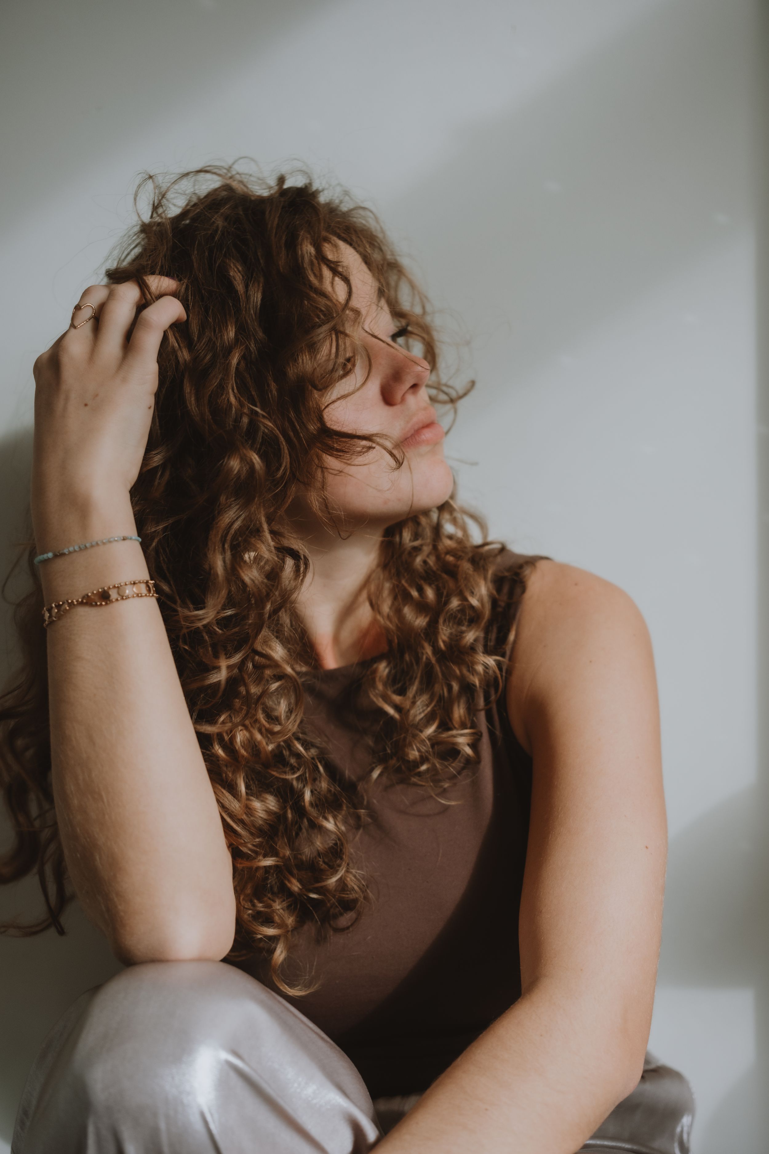 Woman with curly auburn hair in a brown top sitting against a white wall touching her hair.