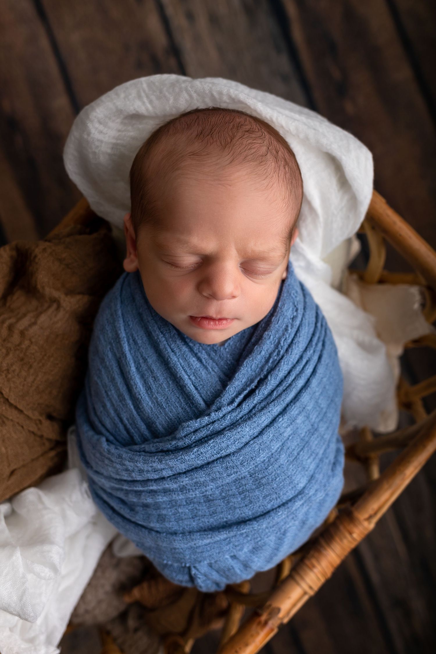 Sleeping newborn wrapped in blue swaddle blanket resting in wooden basket.