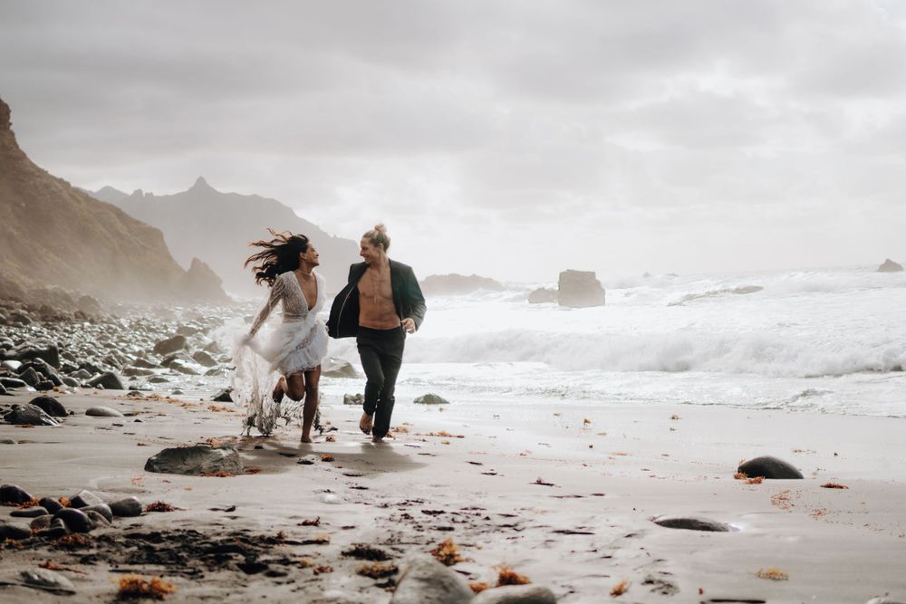 Couple running along a dramatic rocky beach with stormy waves and cliffs.