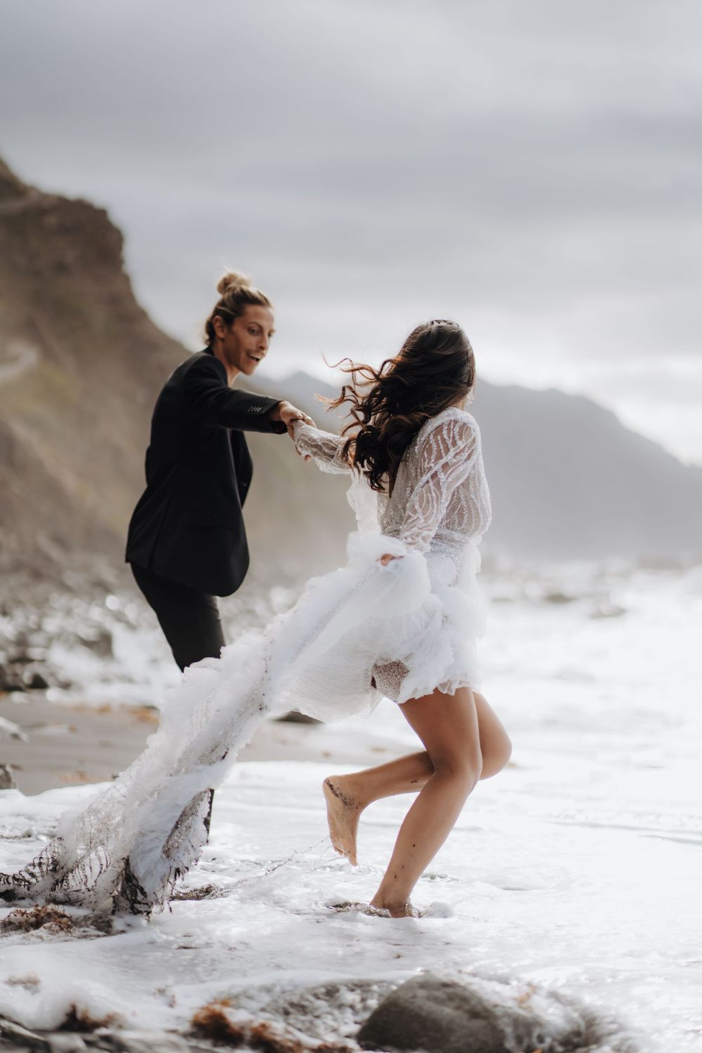 Wind-swept couple playing in the ocean waves on a moody rocky beach.