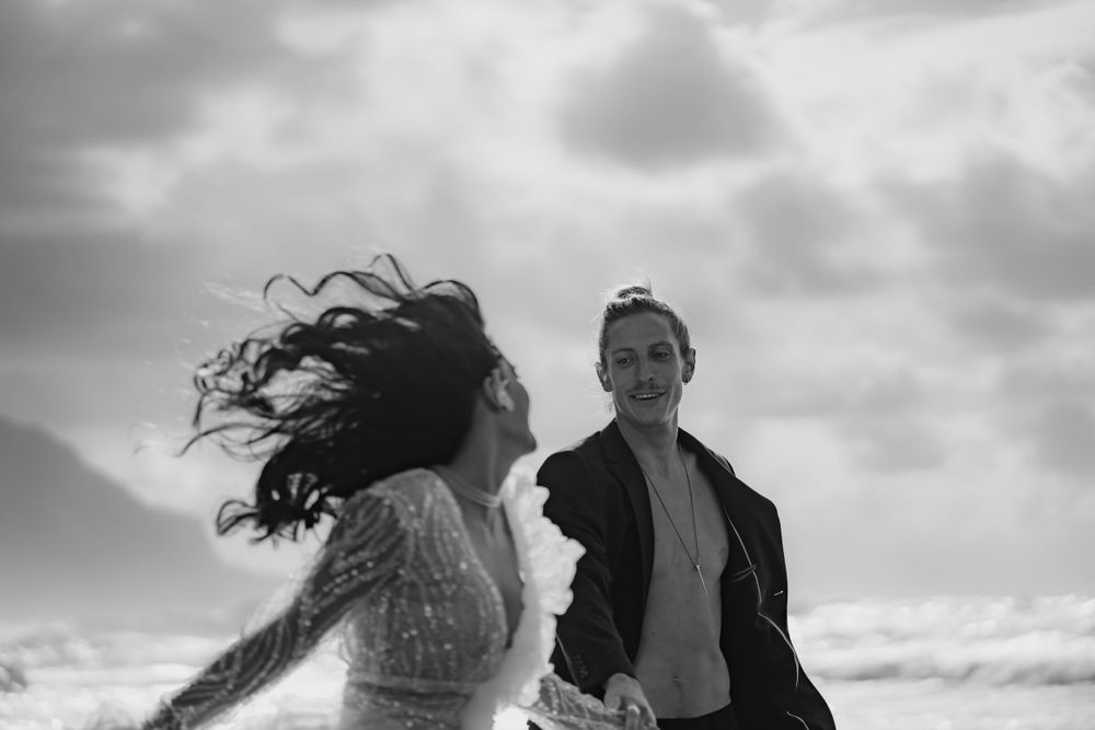 A couple shares a joyful moment on a windy beach with dramatic waves and cloudy skies in the background.