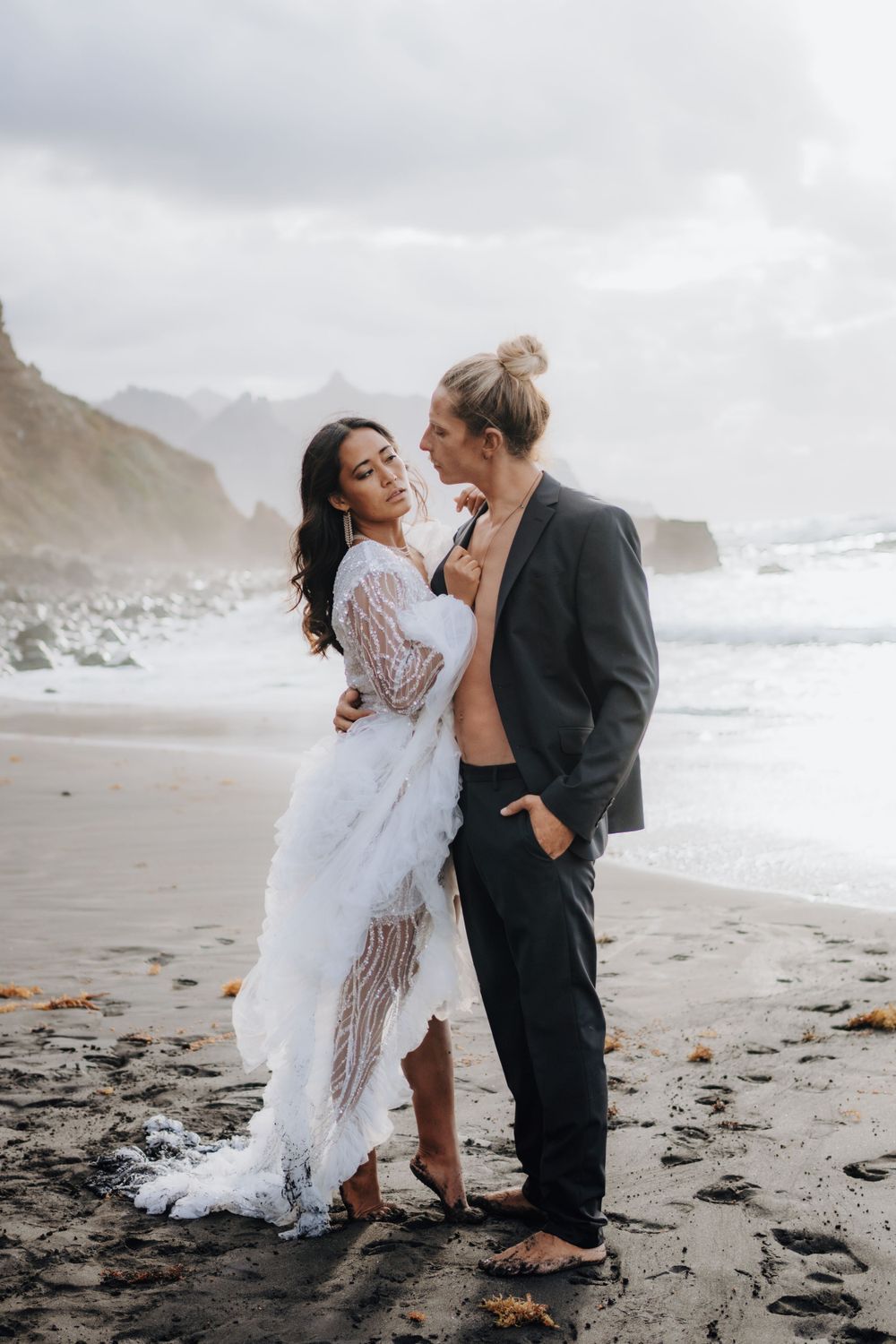 A couple embraces on a rocky beach wearing formal attire against a backdrop of misty cliffs.