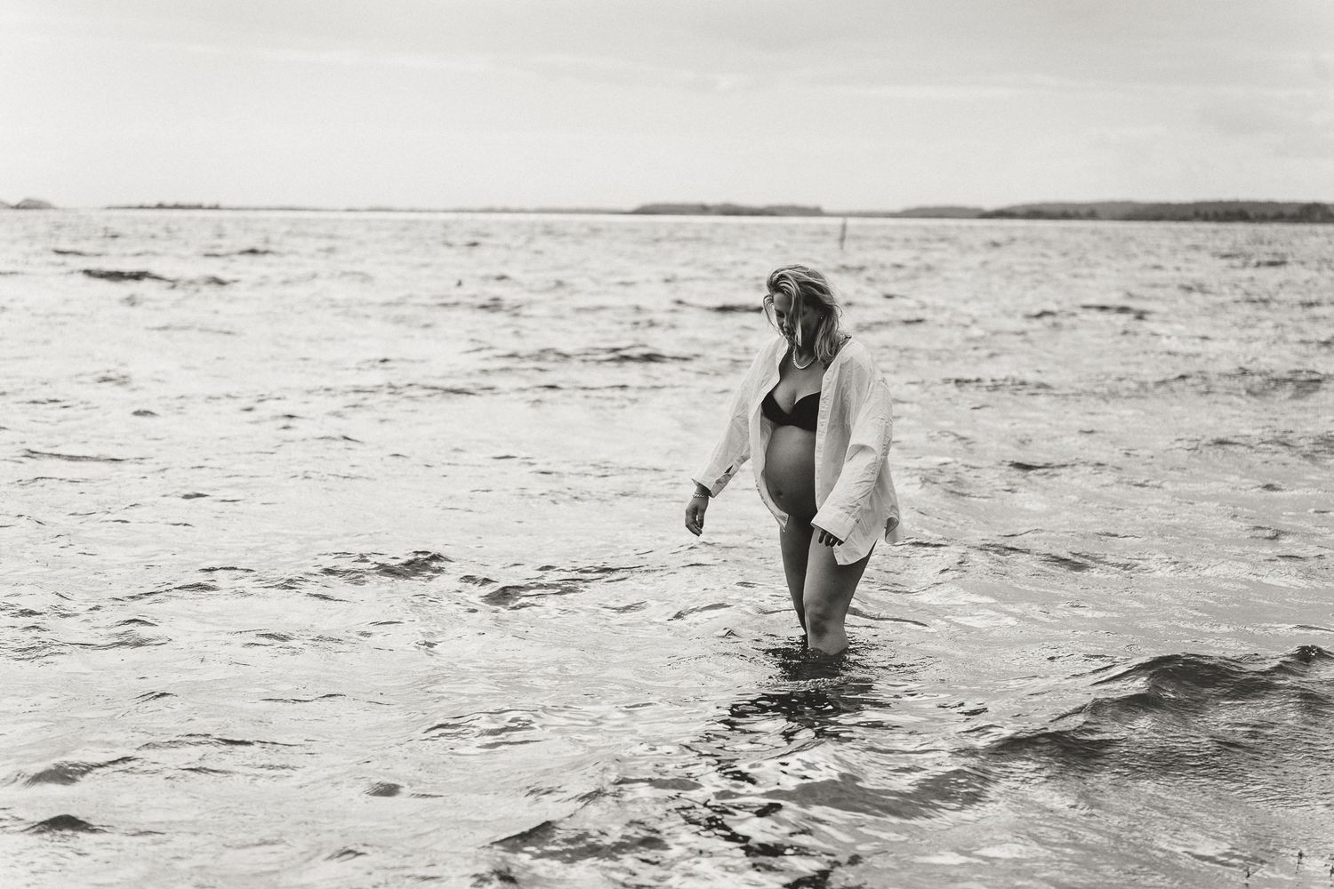 A figure in swimwear and white shirt stands in shallow ocean water on a cloudy day.