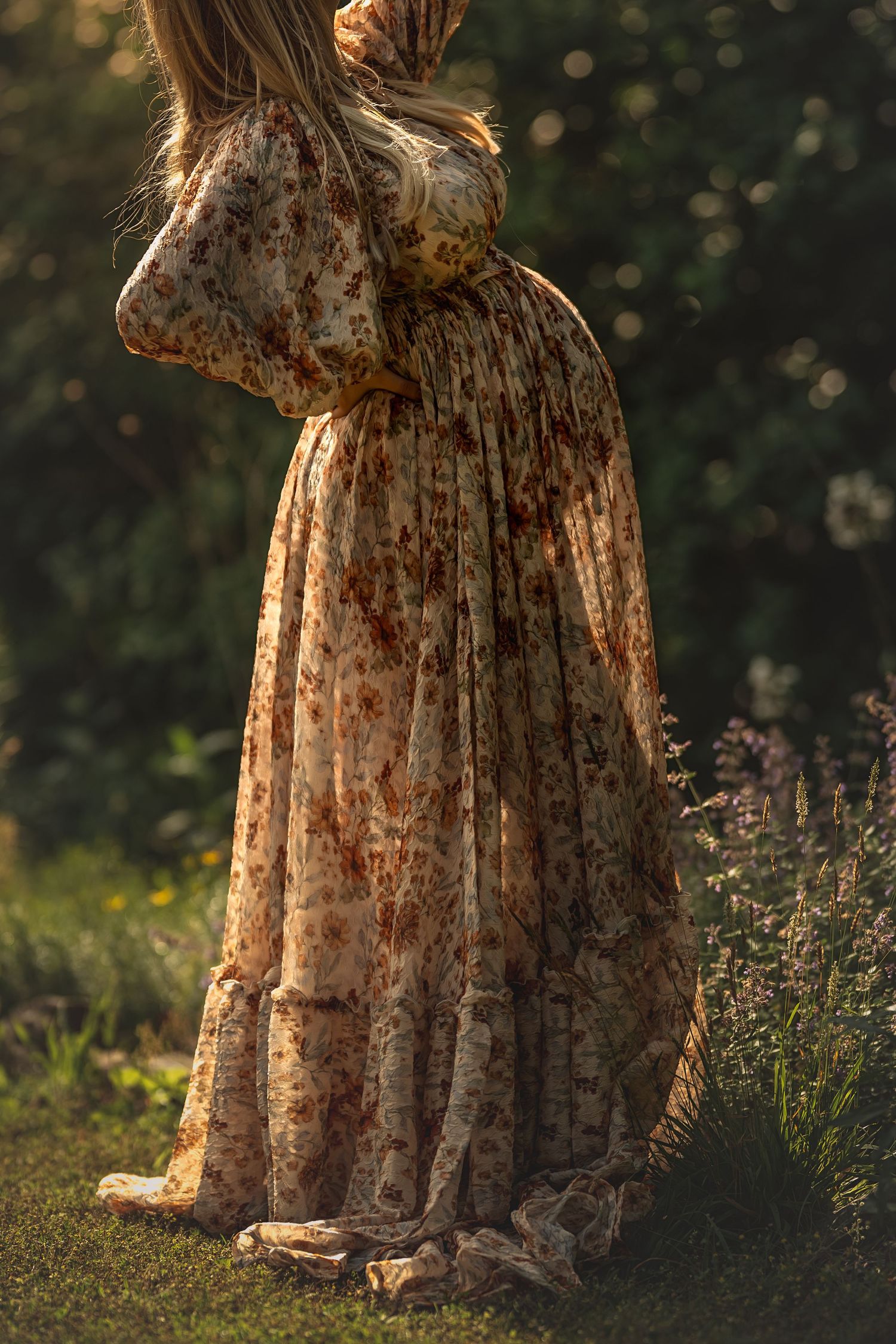 pregnant form visible through sheer floral fabric backlit by golden son in wildflower garden.