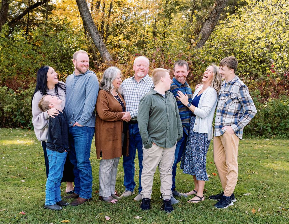 Multi-generational family of eight smiling together outdoors in autumn park setting with colorful fall foliage background.